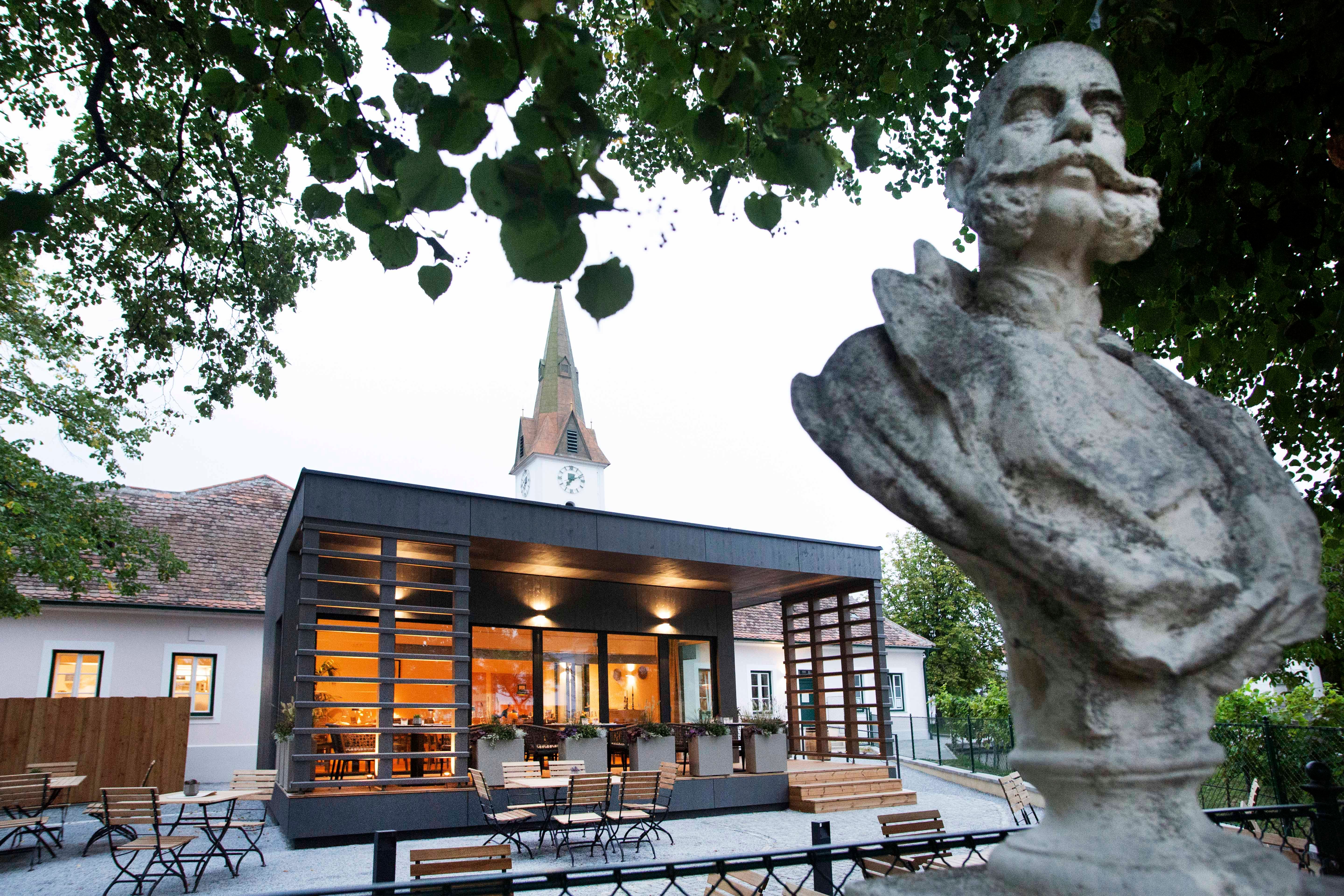 Mellszobor egy modern épület előtt, háttérben egy templomtoronnyal, fákkal körülvéve.