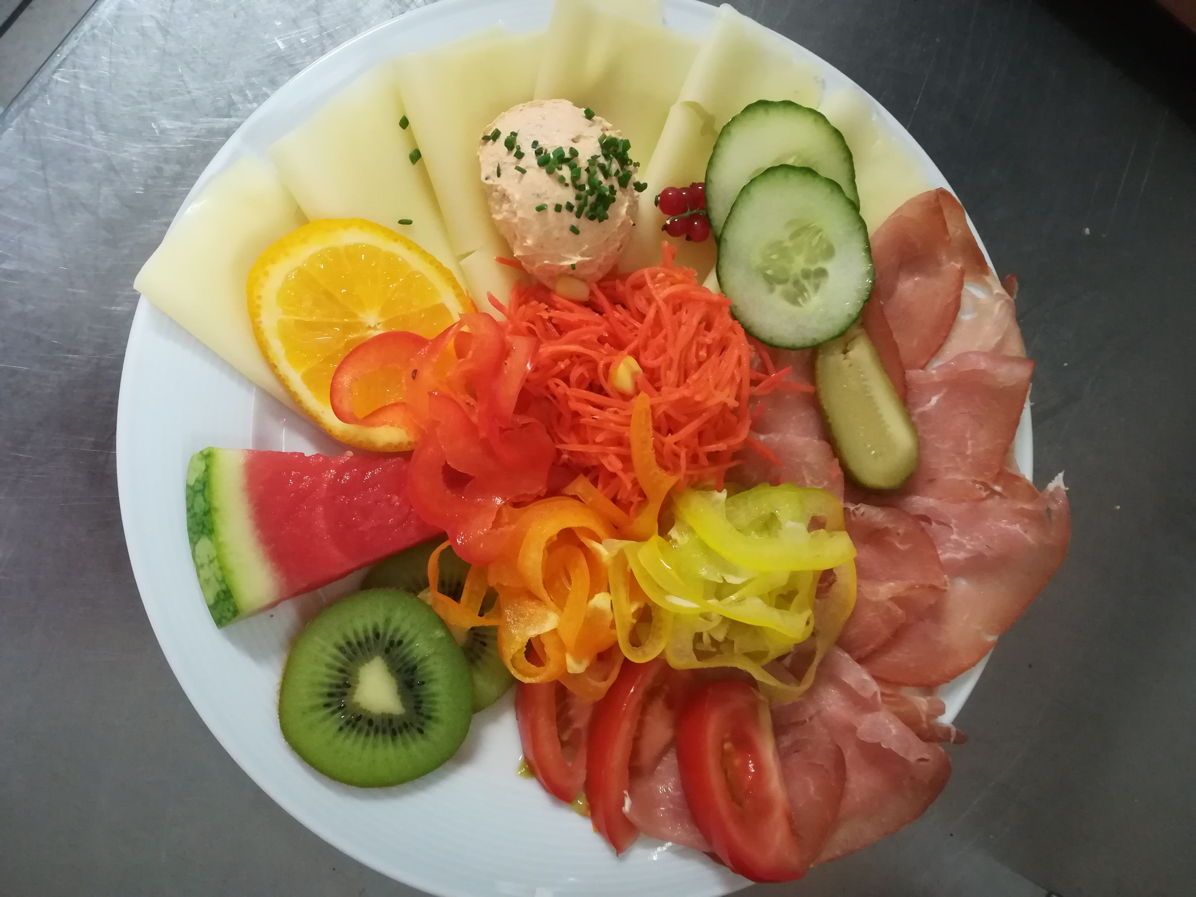 Egy tányér sajt, sonka, zöldségek és gyümölcsök, többek között dinnye, kivi, uborka és paprika.