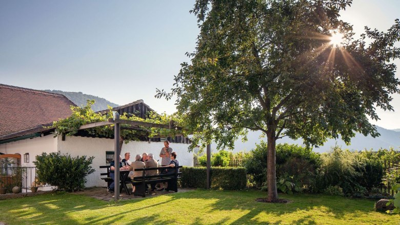 Az emberek a kertben, egy pergola alatt &uuml;lnek egy asztaln&aacute;l, k&ouml;r&uuml;lv&eacute;ve a term&eacute;szettel &eacute;s a naps&uuml;t&eacute;ssel.