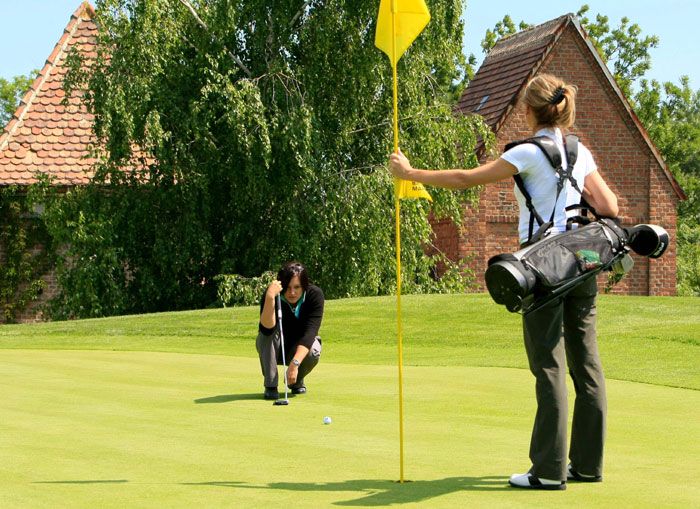 Két nő golfozik egy zöld golfpályán, egyikük a zászlót tartja, a másik a lyukat célozza.