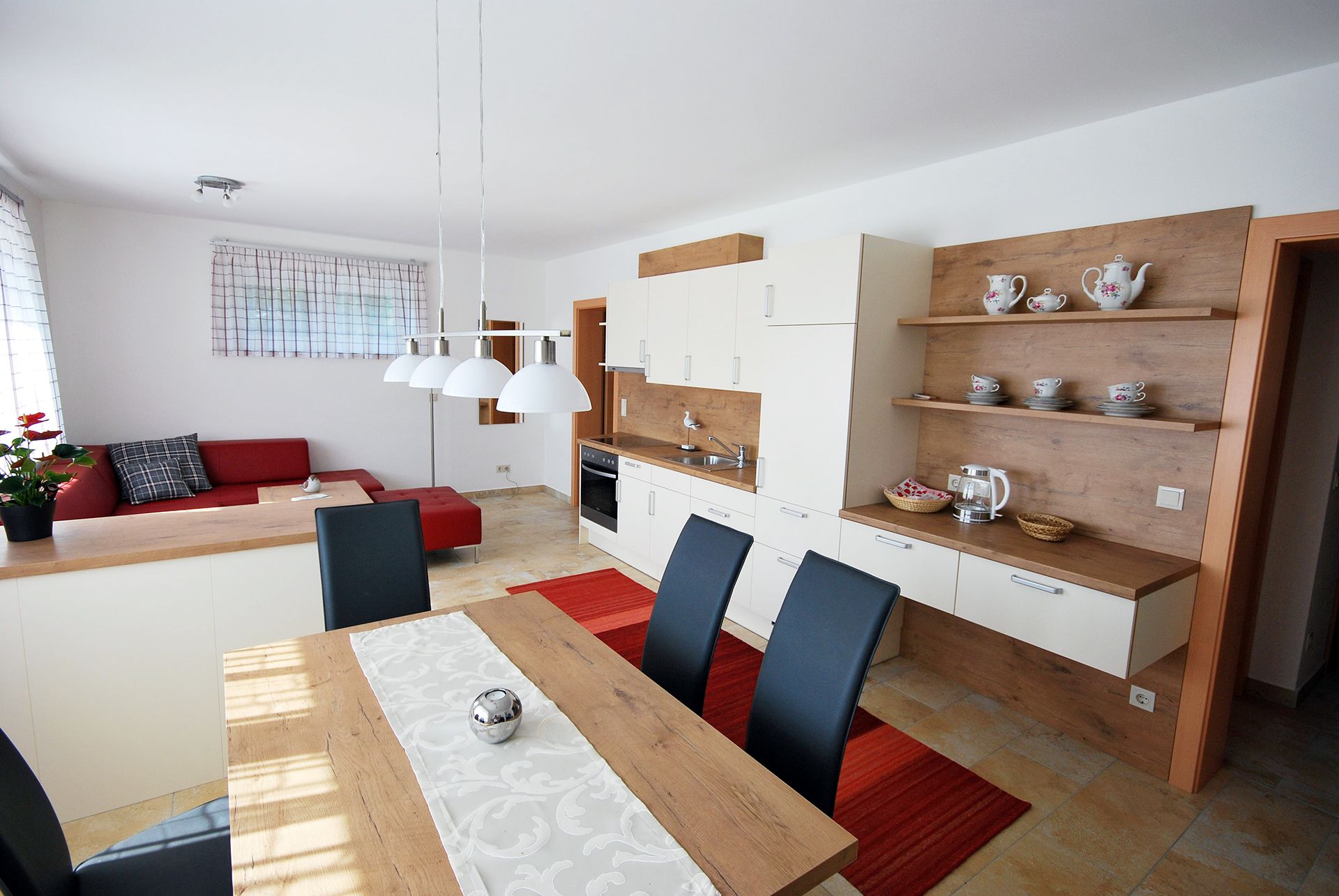 Modern nappali és étkező nyitott konyhával, piros kanapéval, fa asztallal és fehér függőlámpákkal.