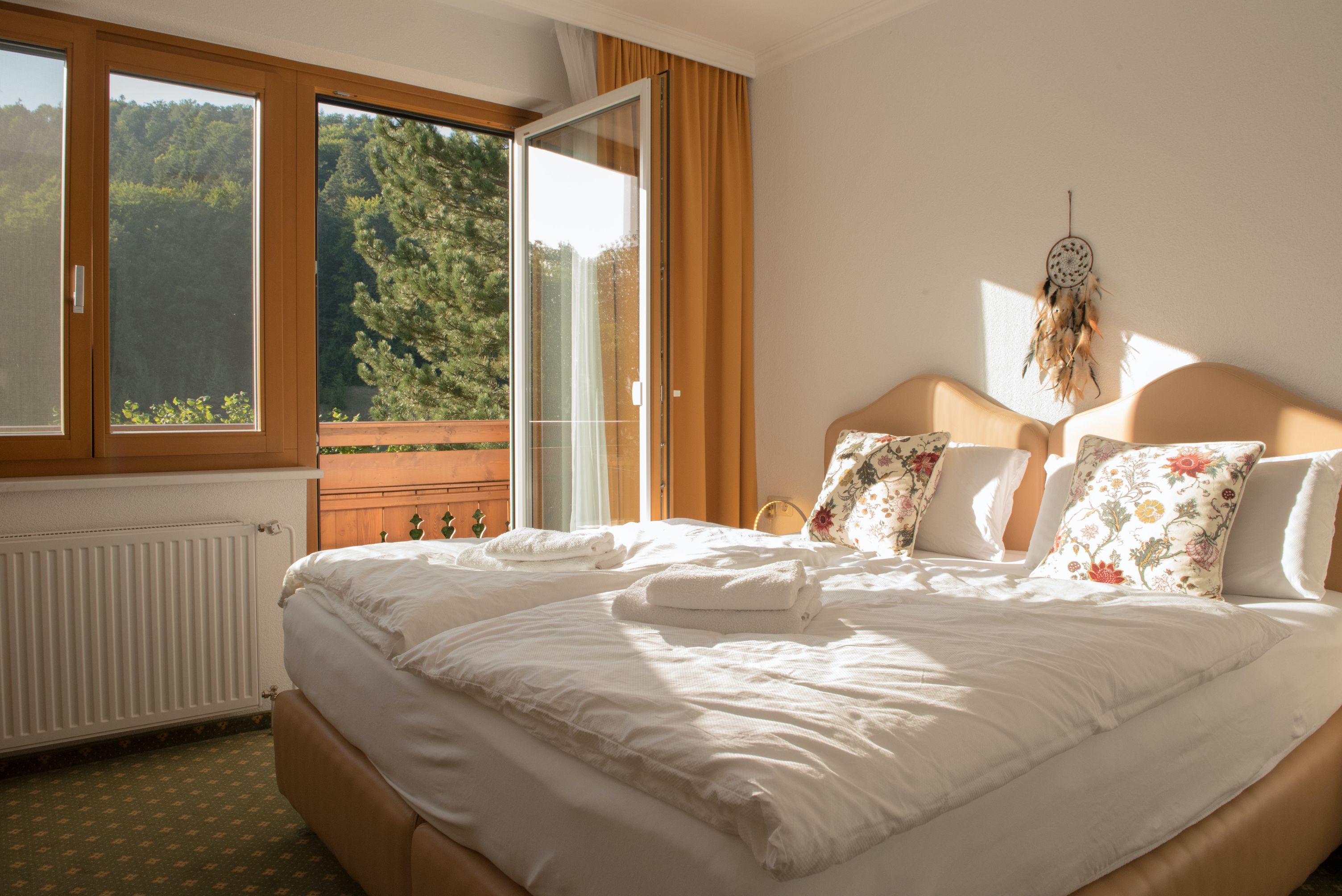 Hangulatos egyágyas szoba franciaággyal, díszpárnákkal és nyitott erkéllyel, kilátással a zöld fákra.