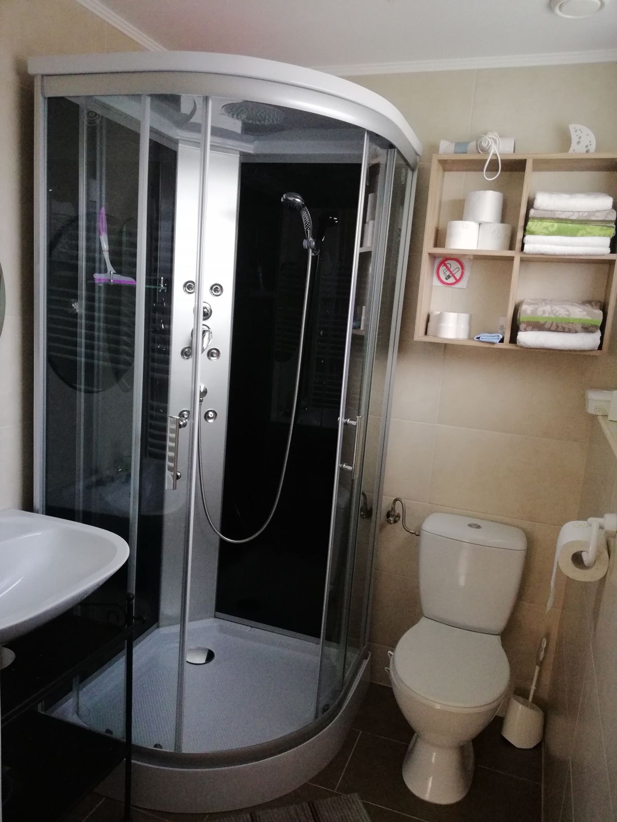 Modern fürdőszoba zuhanyfülkével, mosdókagylóval, WC-vel és polccal, ahol törölköző és WC-papír található.