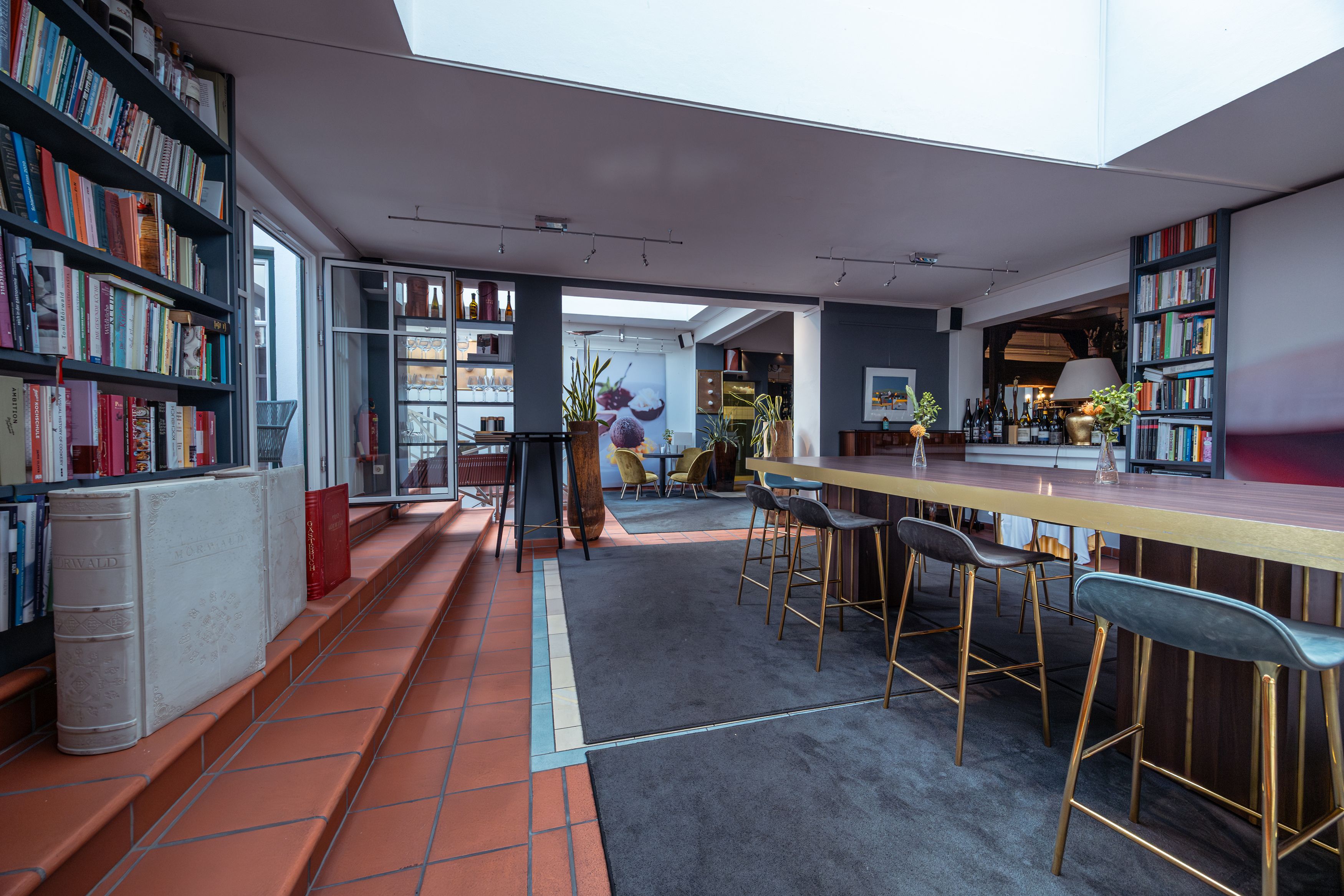 Egy modern szállodai szoba belső nézete könyvespolcokkal, magas asztalokkal és bárszékekkel.