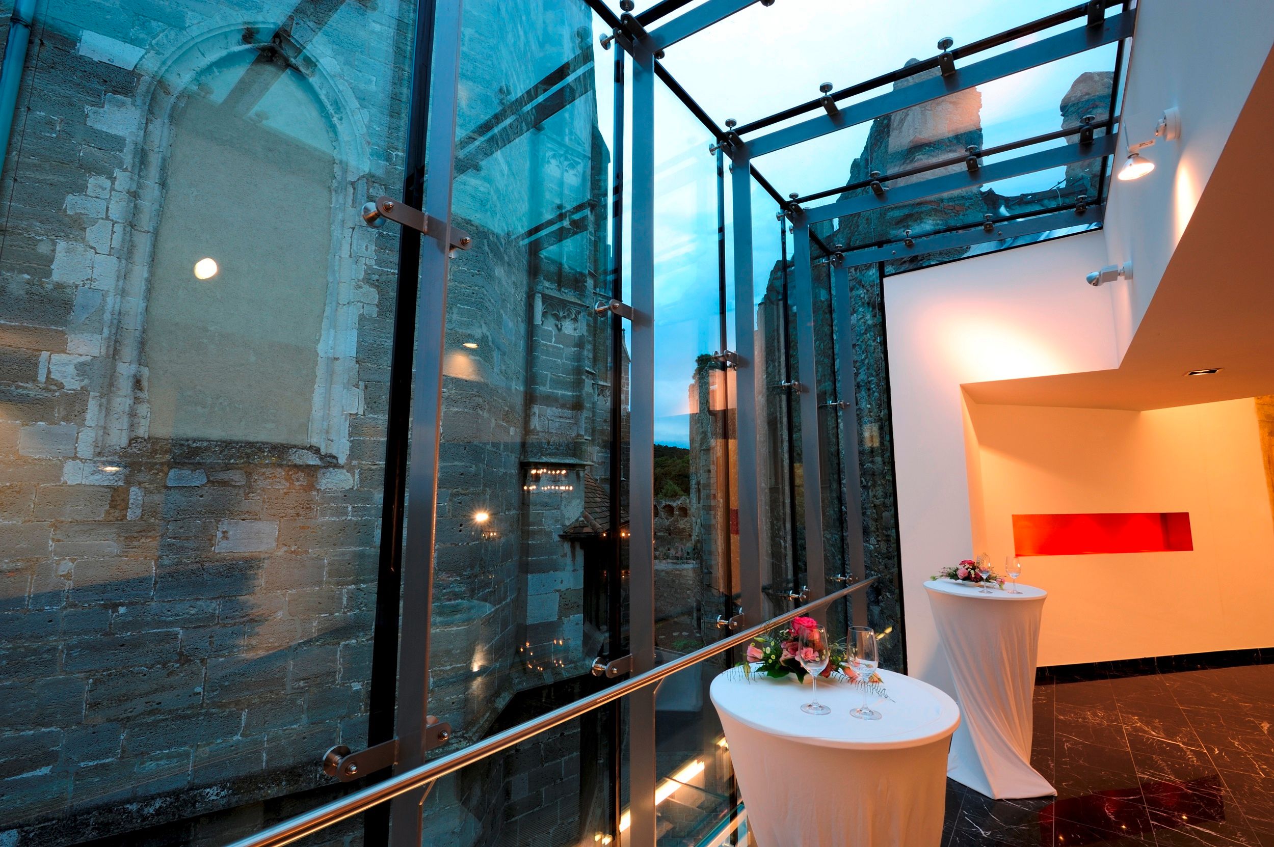 Modern üvegépítészet egy régi kőfal mellett, díszített bárasztalokkal.