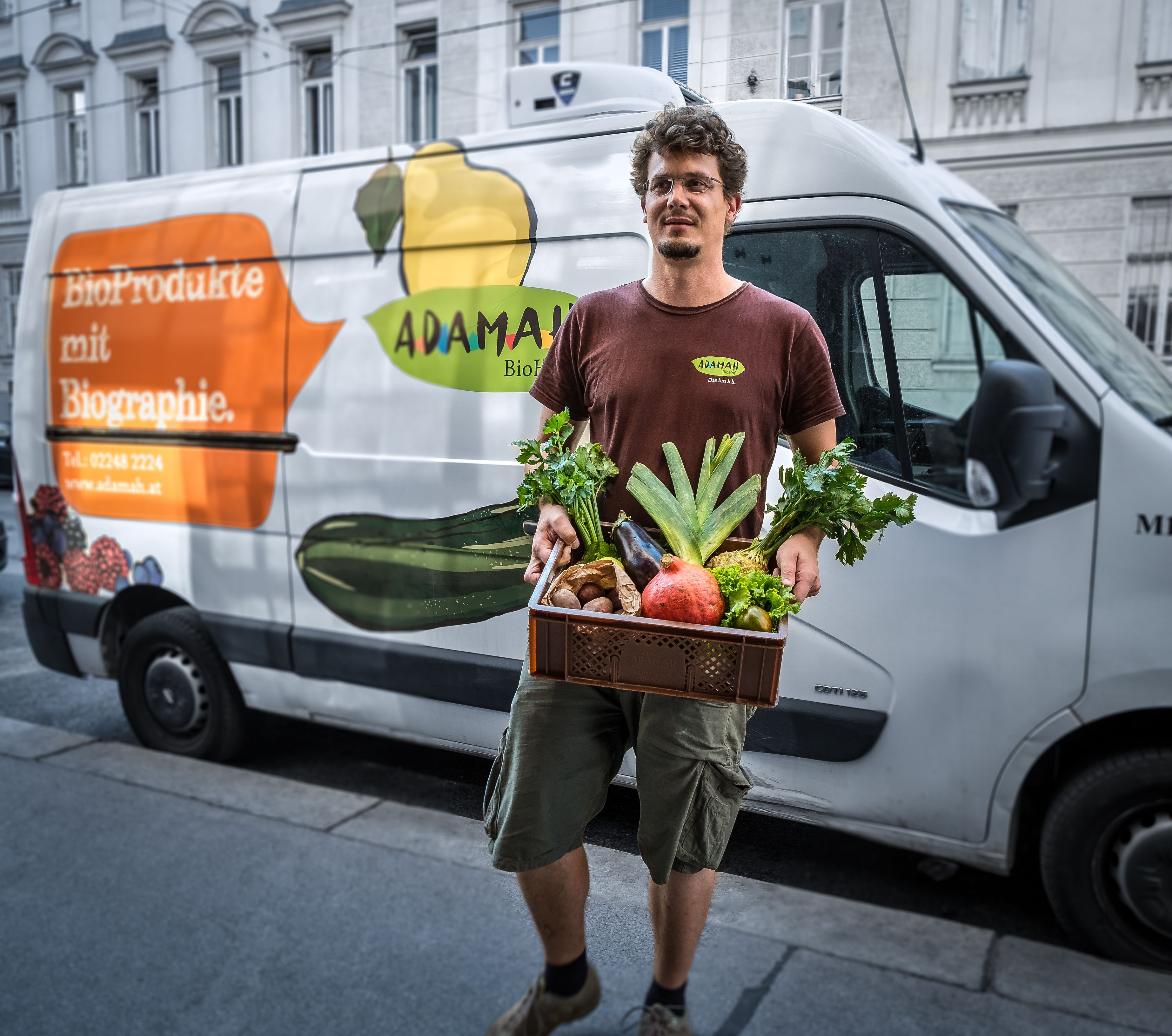 Egy férfi egy doboz friss gyümölcsöt és zöldséget tart egy ADAMAH-logóval ellátott áruszállító furgon előtt.