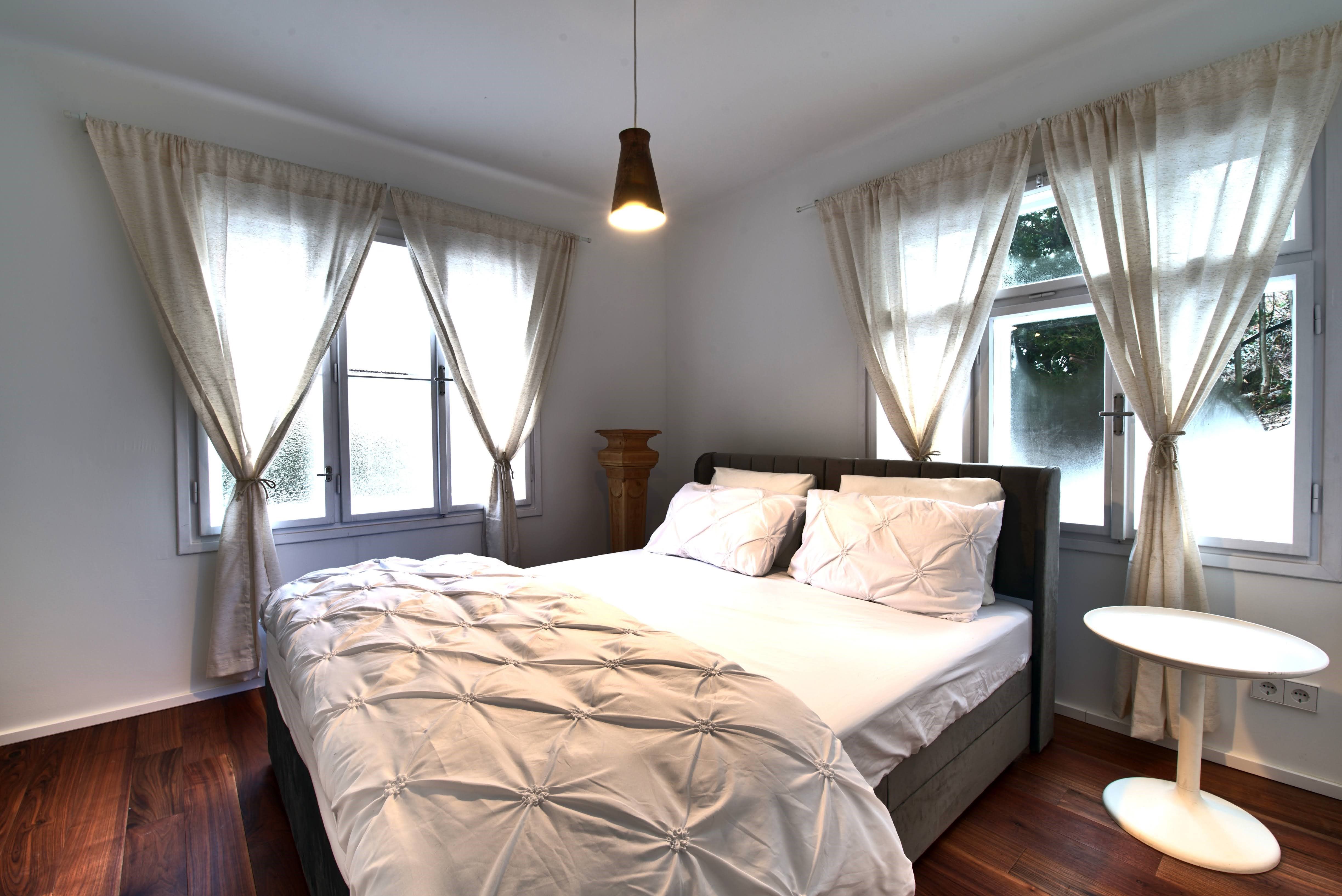 Hangulatos hálószoba nagy ággyal, fehér függönyökkel és egy kis kerek asztallal.