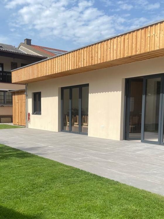 Modern épület külső fa burkolattal és nagy ablakokkal, terasszal és pázsittal körülvéve.