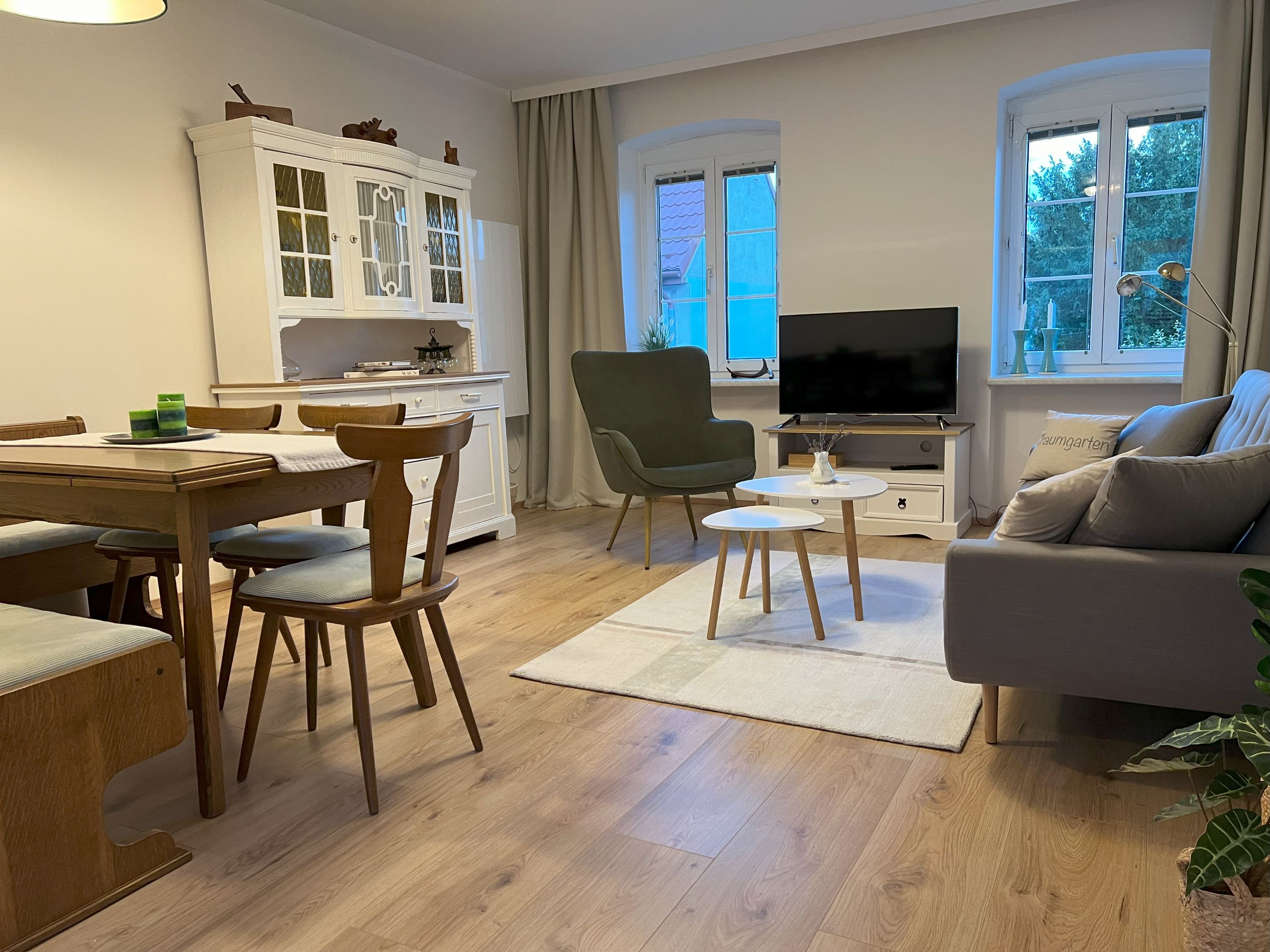 Hangulatos nappali fabútorokkal, kanapéval, fotelekkel, étkezőasztallal és TV-vel. Világos falak és fapadló.