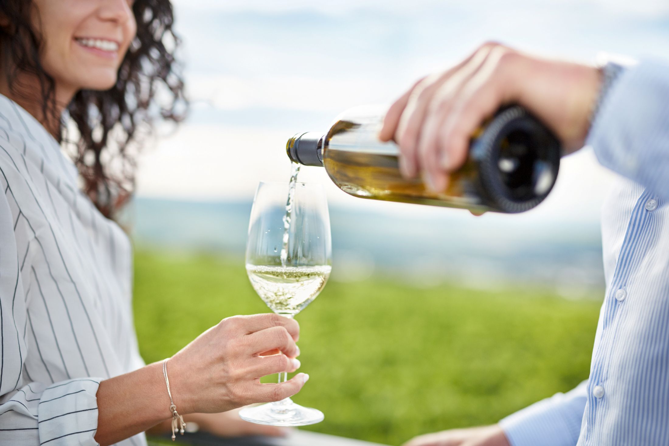 Egy személy fehérbort tölt egy másik személy által tartott pohárba, a háttérben egy szőlőskert.