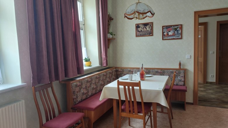 Hangulatos &eacute;tkezős konyha &eacute;tkezőasztallal, sz&eacute;kekkel &eacute;s sarokpaddal. Ablak f&uuml;gg&ouml;ny&ouml;kkel, k&eacute;pek a falon.