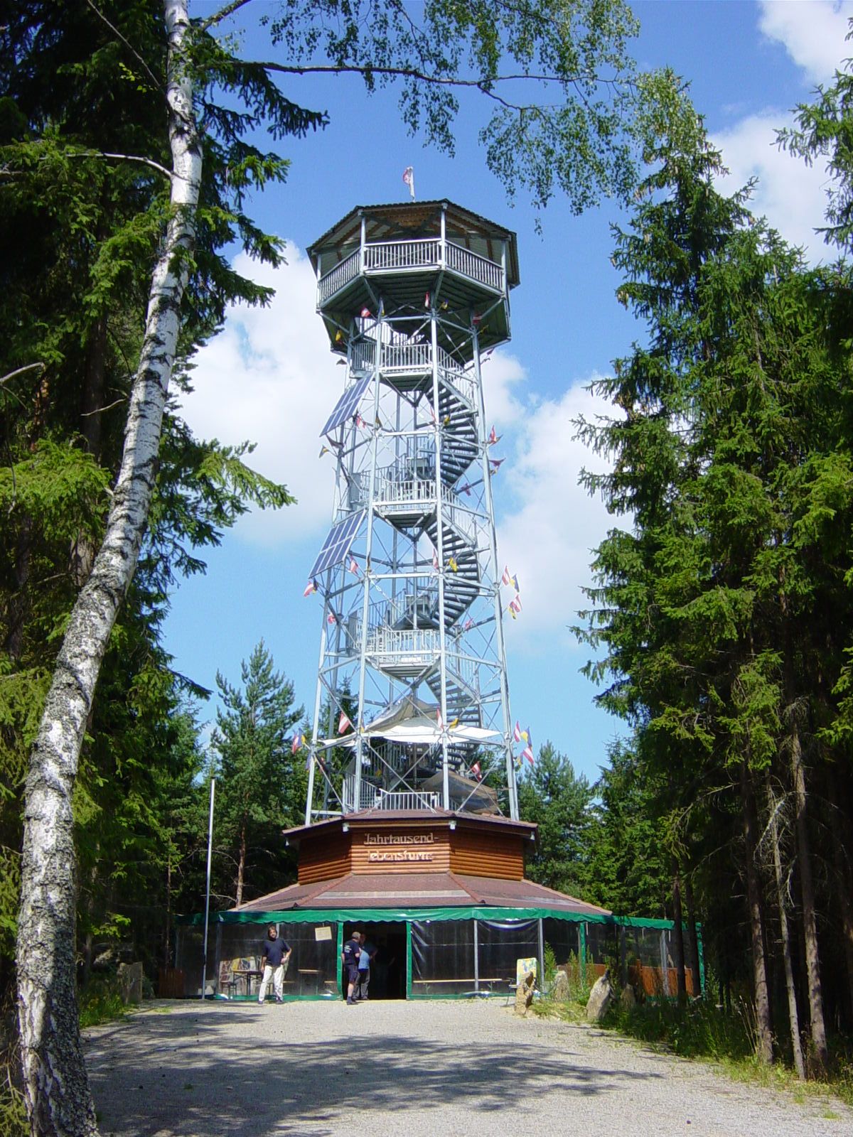 Magas, fából készült, fával burkolt fém kilátótorony, fákkal körülvéve, kék ég alatt.