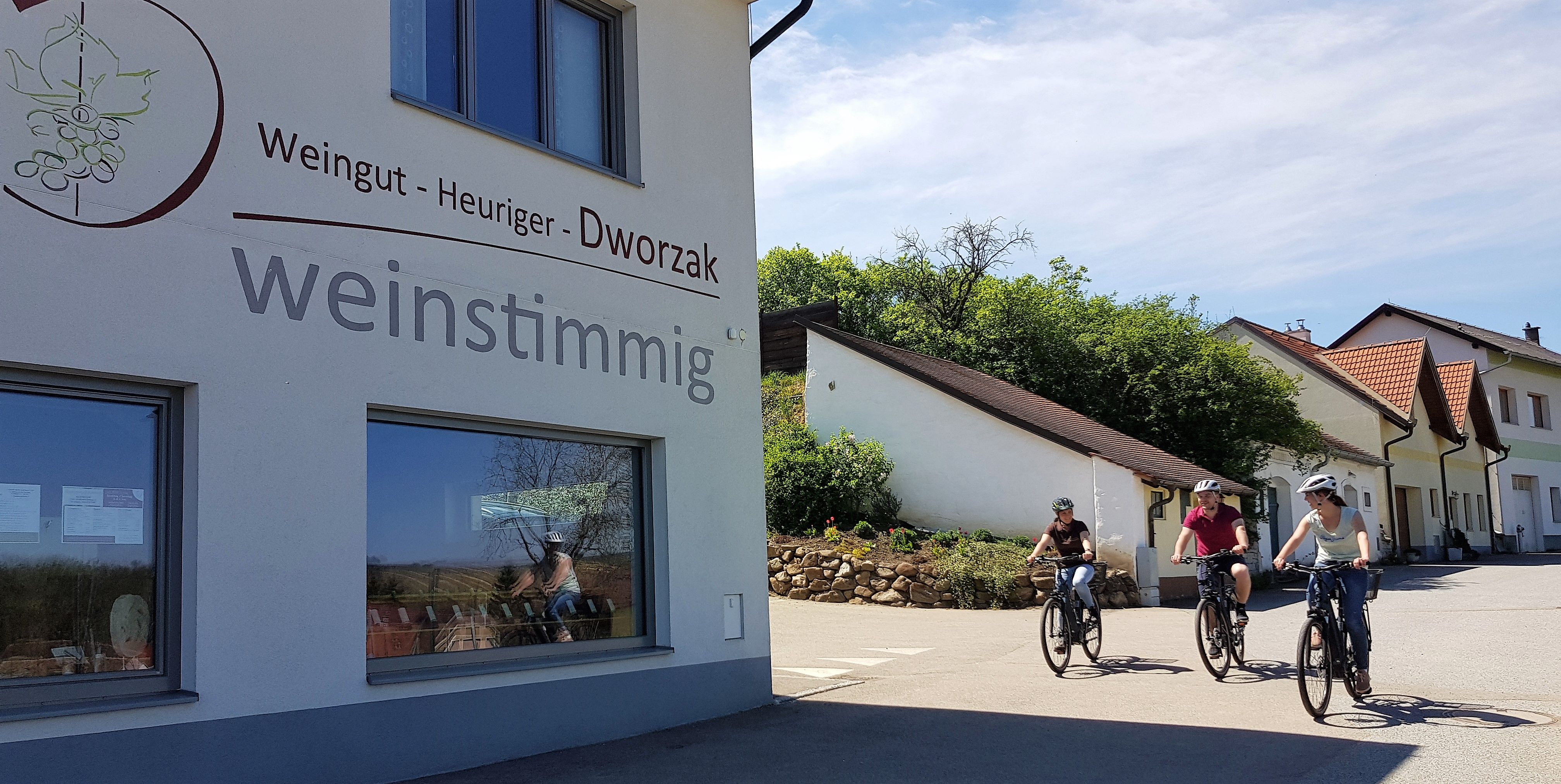 Három kerékpáros halad el egy "Weingut - Hagyományos borozó - Dworzak weinstimmig" feliratú épület mellett.
