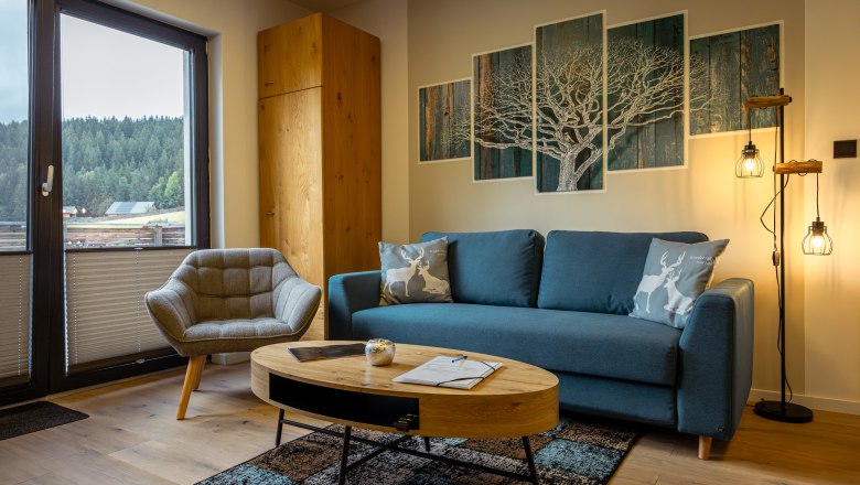 Hangulatos nappali kék kanapéval, karosszékkel, fa asztallal és az ablakon keresztül erdei kilátással.