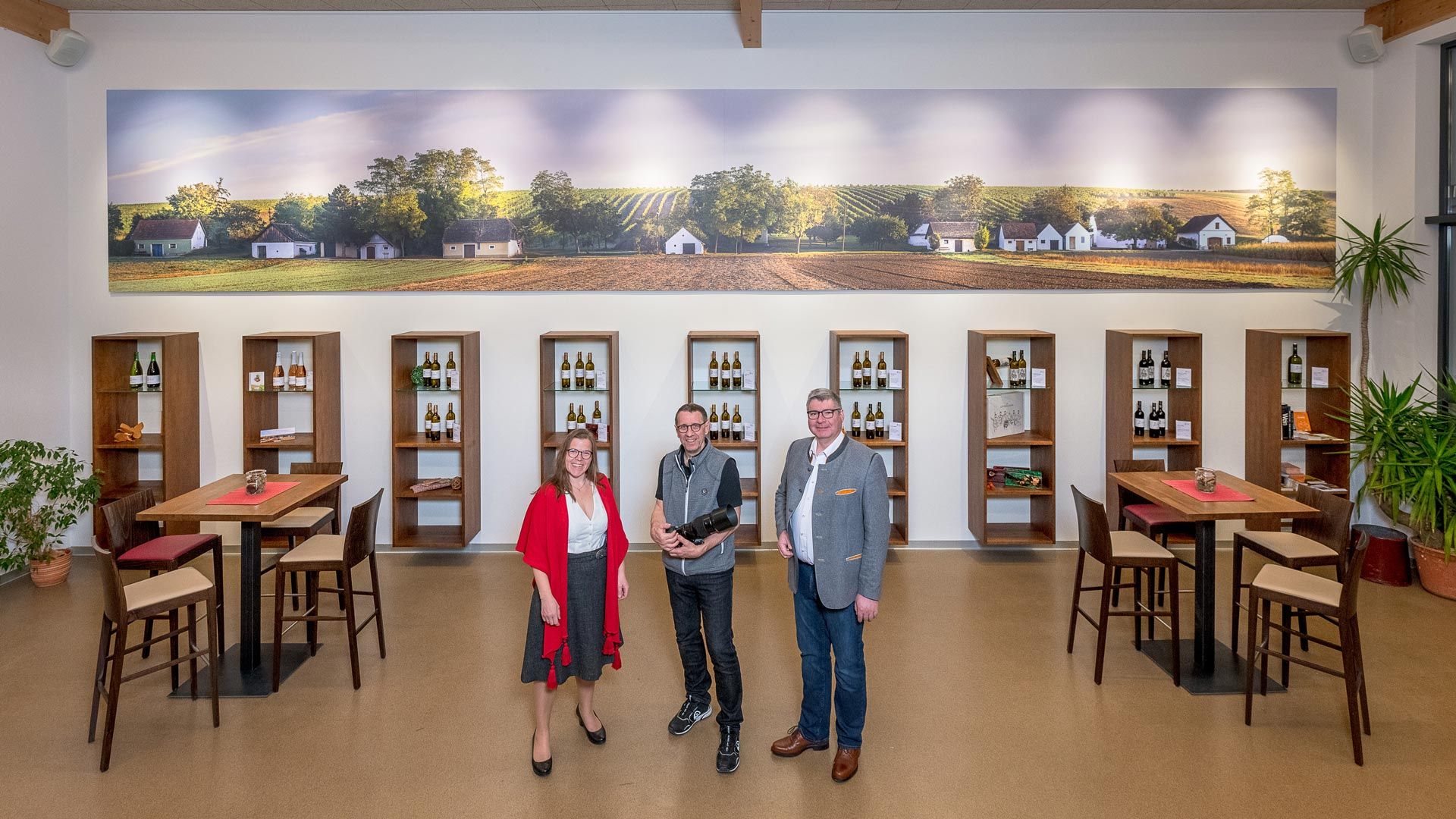 Három ember áll egy borászatban, borosüvegek polcai és egy nagy, szőlőültetvényeket ábrázoló falfestmény előtt.