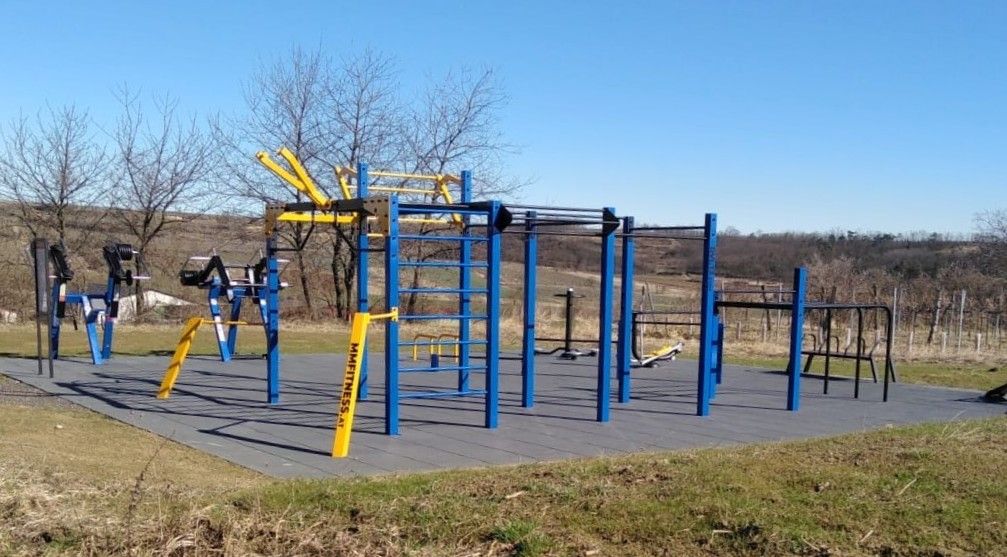 Szabadtéri kaliszténika-park kék és sárga eszközökkel, vidéki tájjal körülvéve.