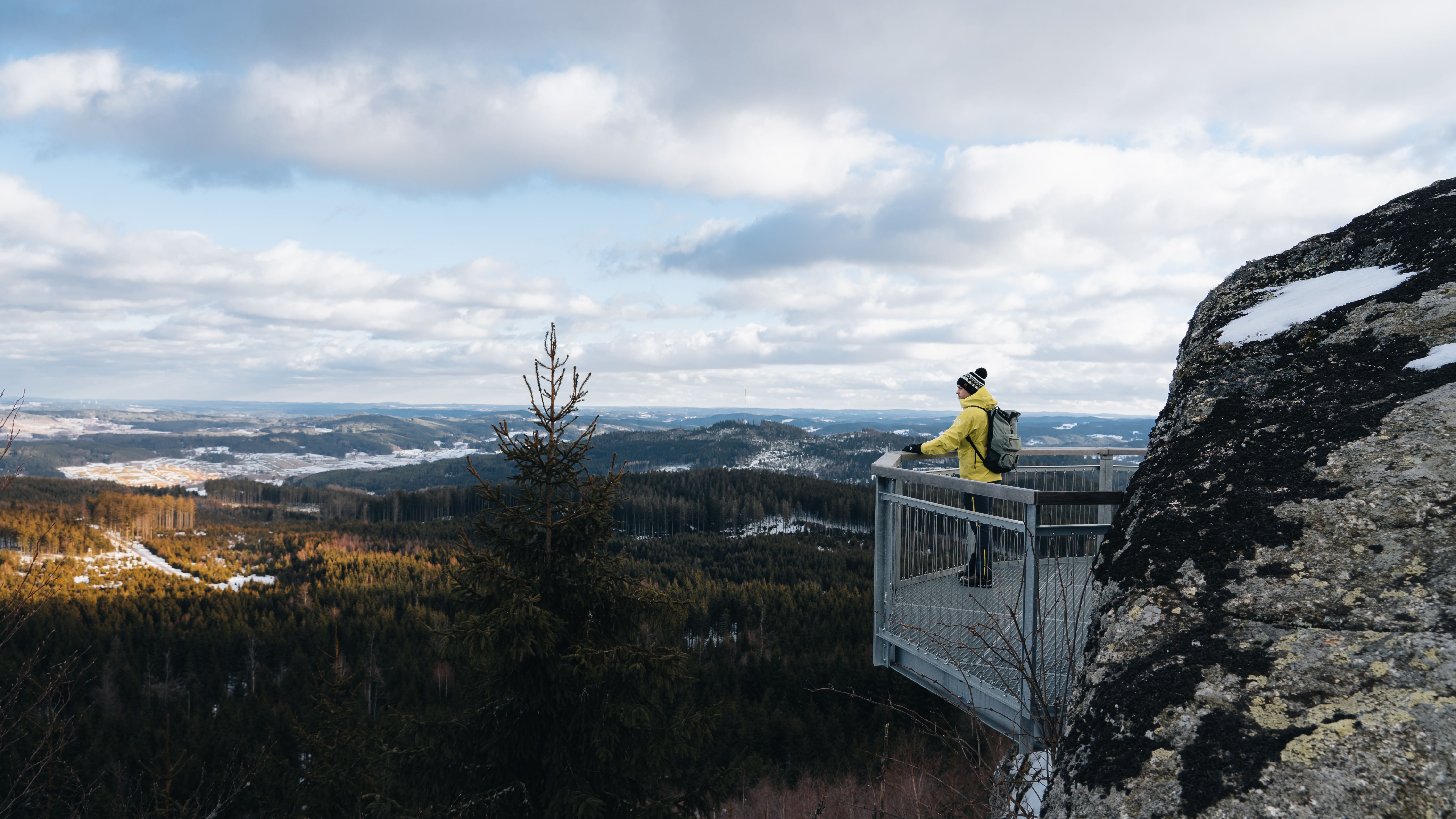 Ein Wanderer genießt die atemberaubende Aussicht auf die verschneiten Wälder und sanften Hügel, die sich bis zum Horizont erstrecken. Die frische, klare Winterluft und die ruhige Atmosphäre laden dazu ein, die Schönheit der Natur in vollen Zügen zu erleben.