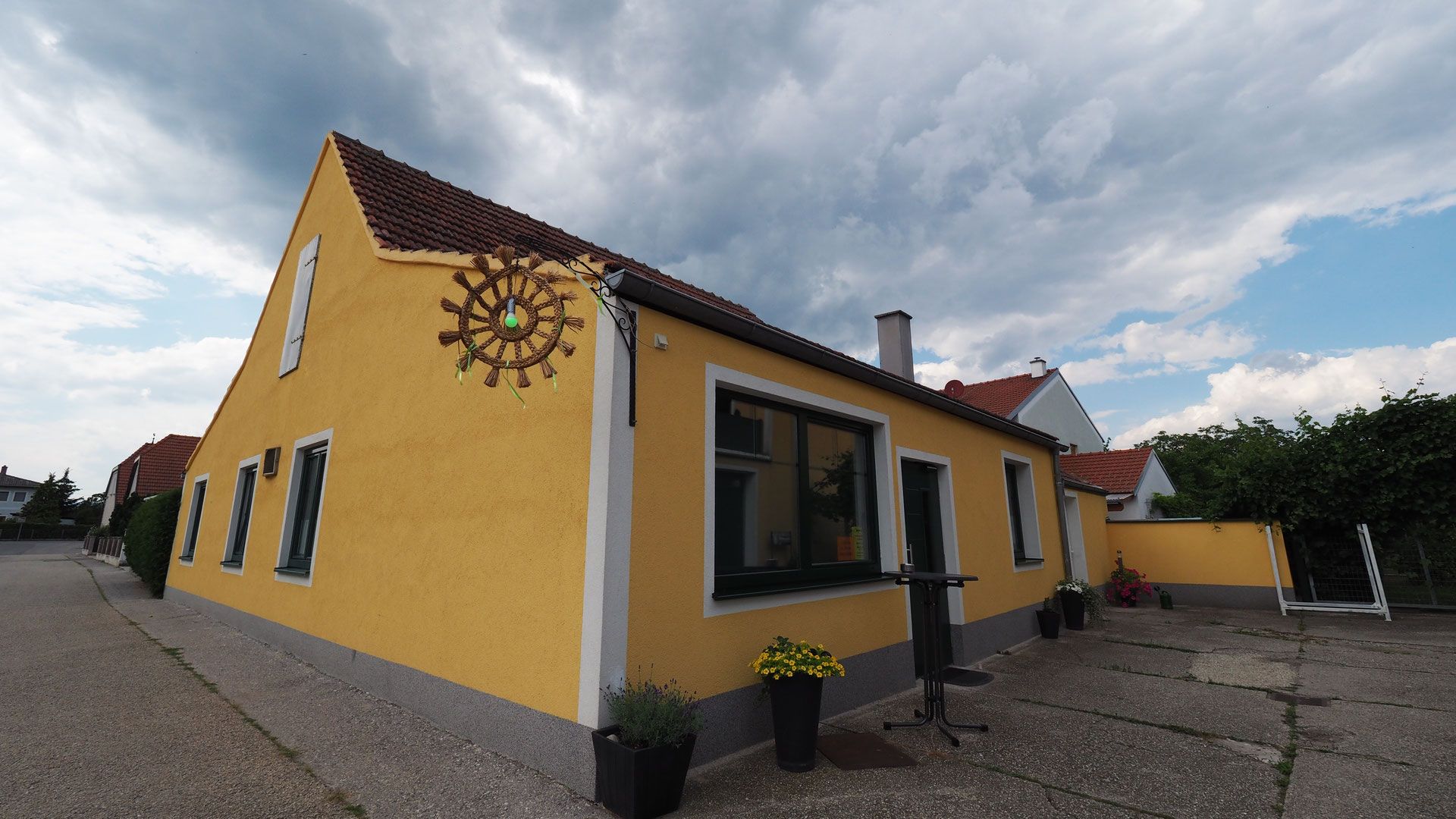Sárga épület piros tetőcserepekkel és egy díszes kerékkel a falon, növényekkel és felhős égboltal körülvéve.