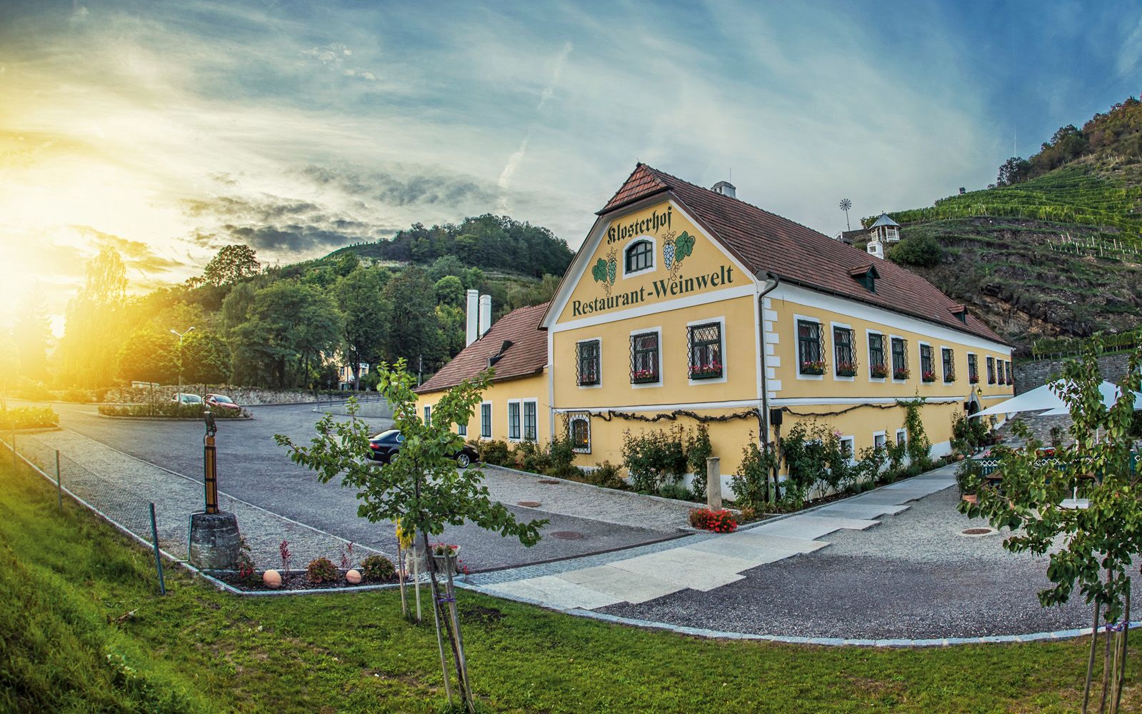A "Klosterhof Restaurant-Weinwelt" feliratú sárga épület külső nézete, fákkal és szőlőültetvényekkel körülvéve, naplementében.