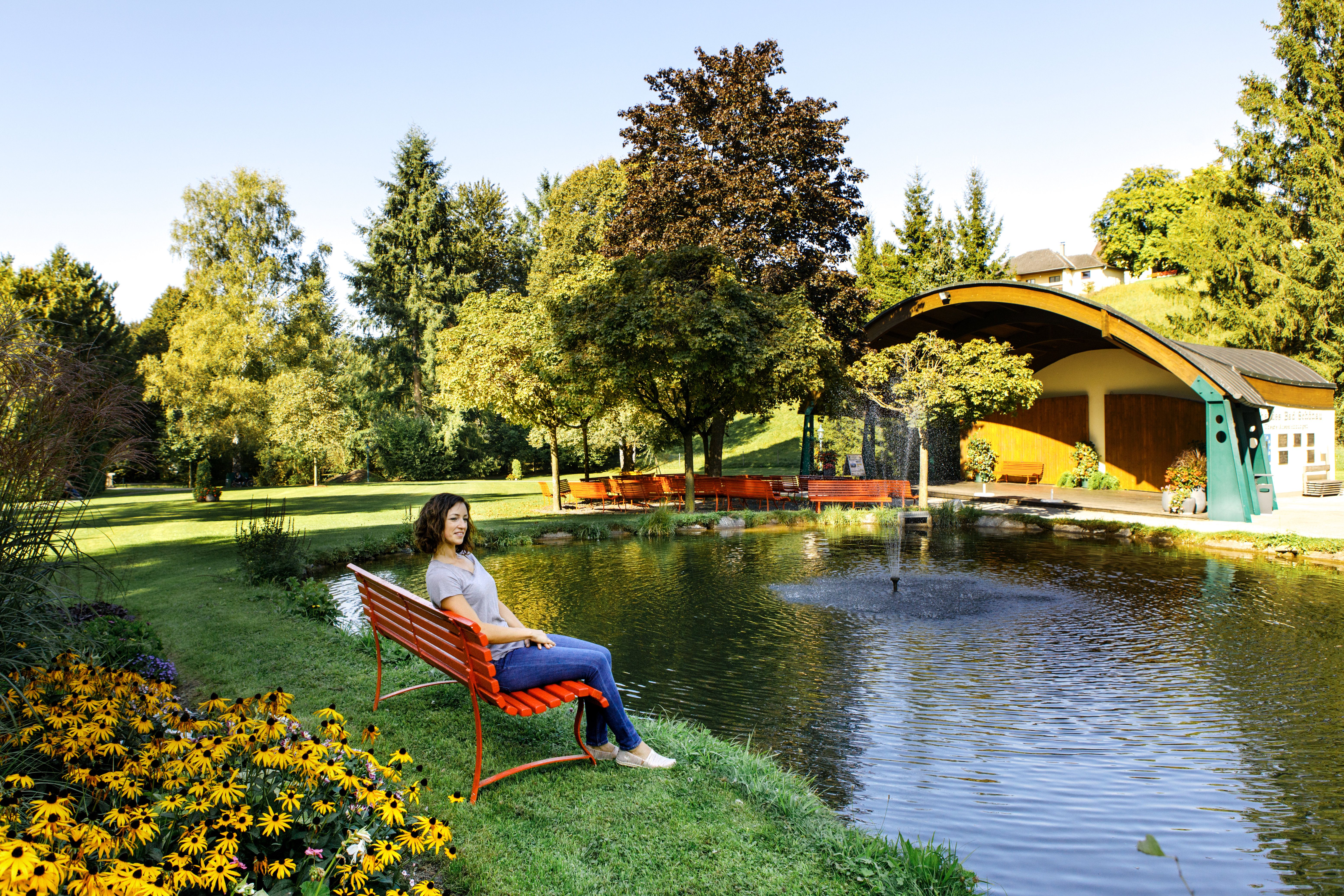 Nő ül egy piros padon a Bad Schönau-i gyógyfürdőkertben lévő tó mellett.
