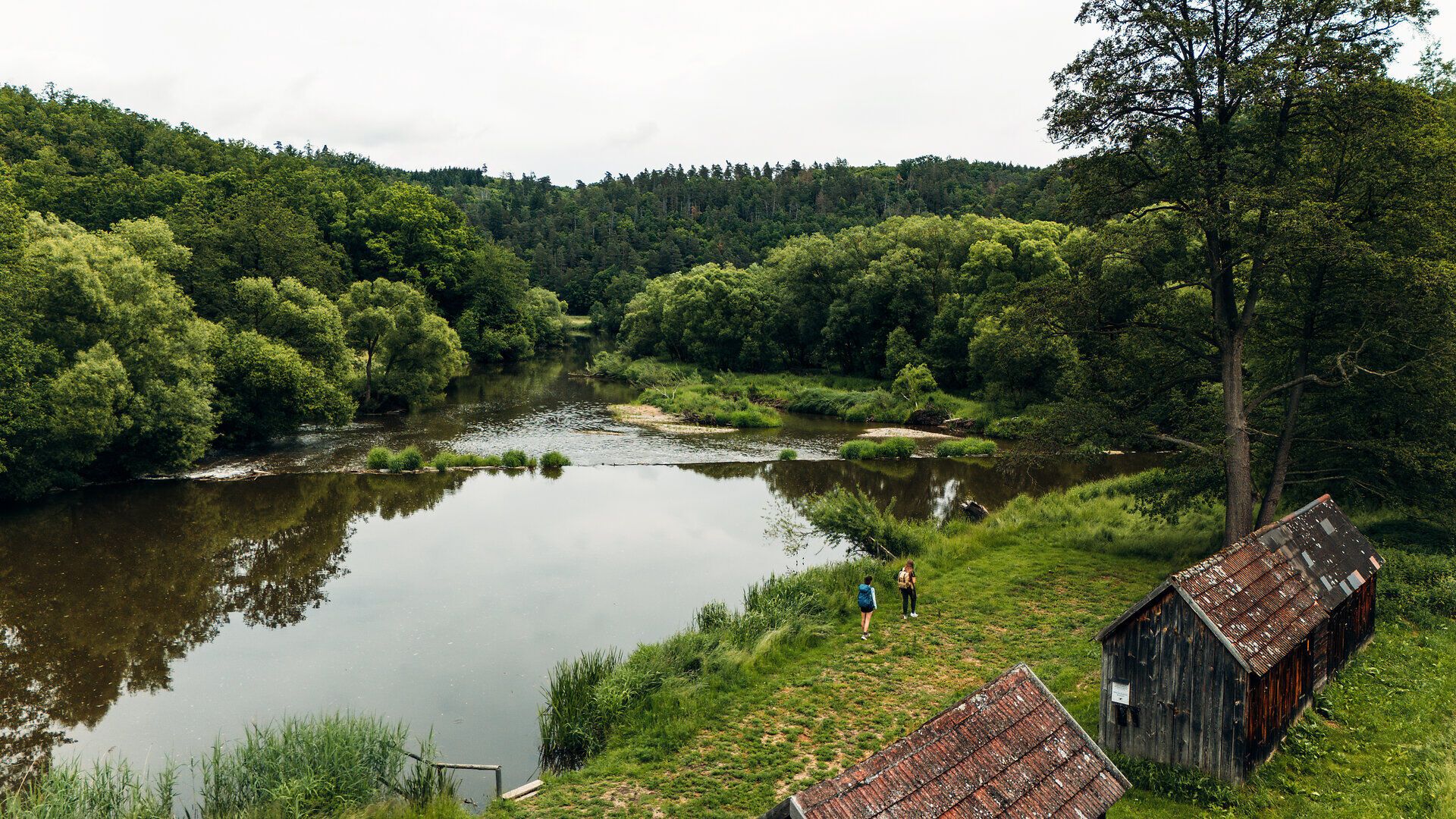 Két ember sétál a sűrű fákkal és régi faházakkal szegélyezett folyóparton a Waldviertelben található Drosendorfban.