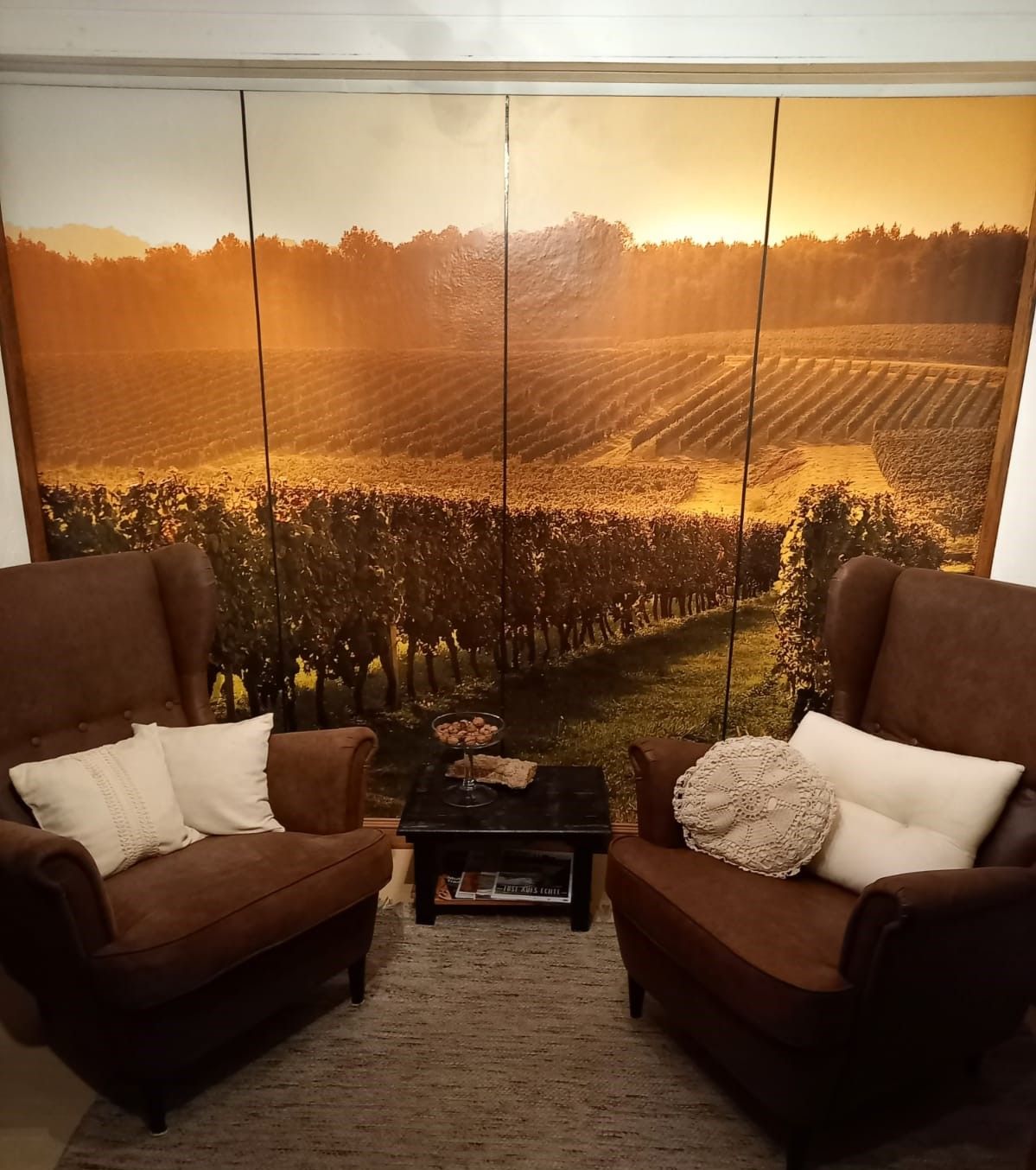 Két barna, párnázott fotel egy fal előtt, amelyen egy szőlőskertet ábrázoló tájképes fotótapéta látható.