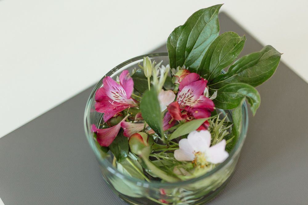 Egy üvegedény színes virágokkal és zöld levelekkel egy asztalon.