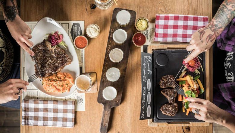 Két ember steaket eszik köretekkel egy faasztalnál.