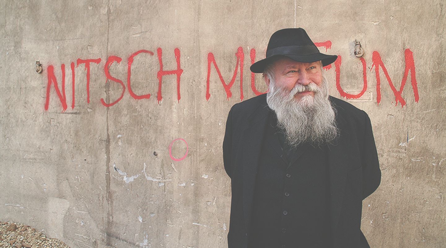 Egy szakállas, kalapos férfi áll a "Nitsch Múzeum" feliratú fal előtt.