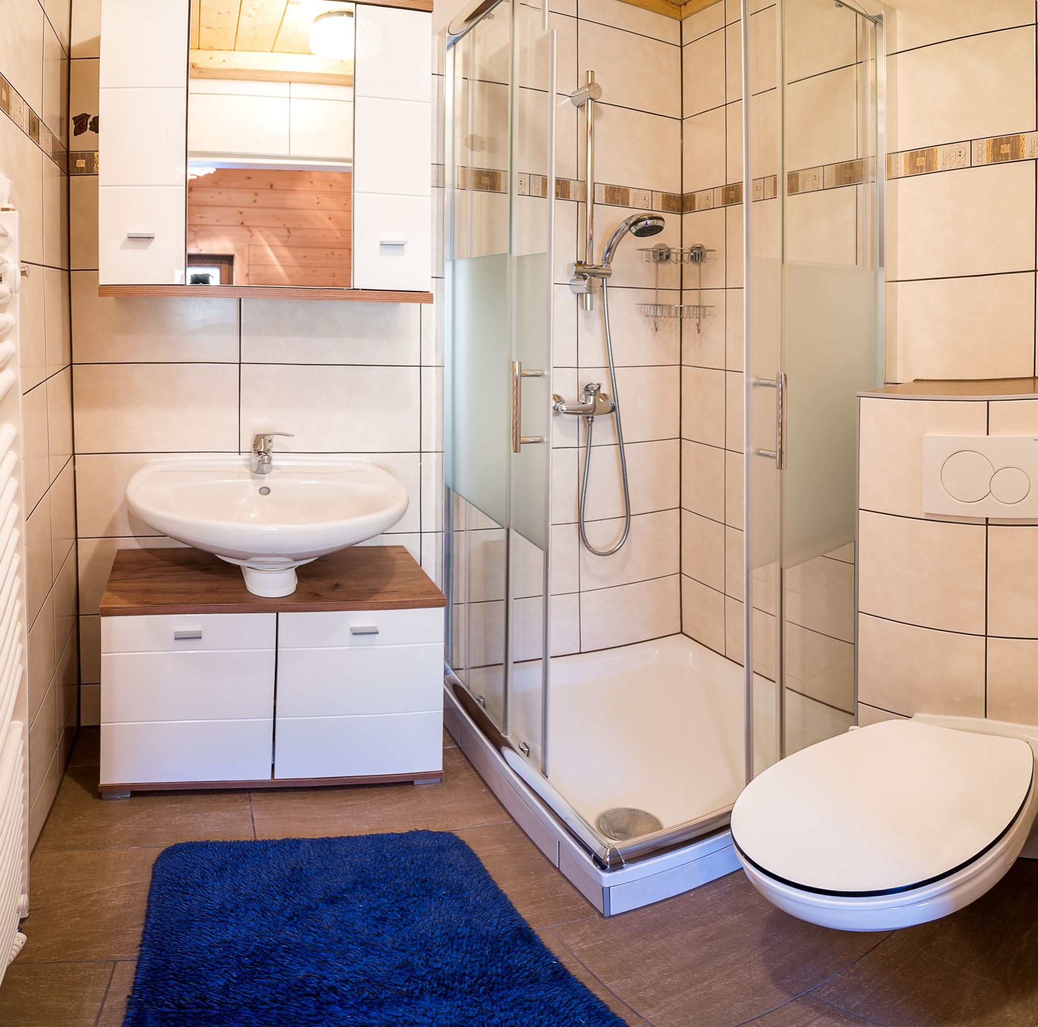 Modern fürdőszoba zuhanyzóval, mosdóval, WC-vel és kék fürdőszőnyeggel.