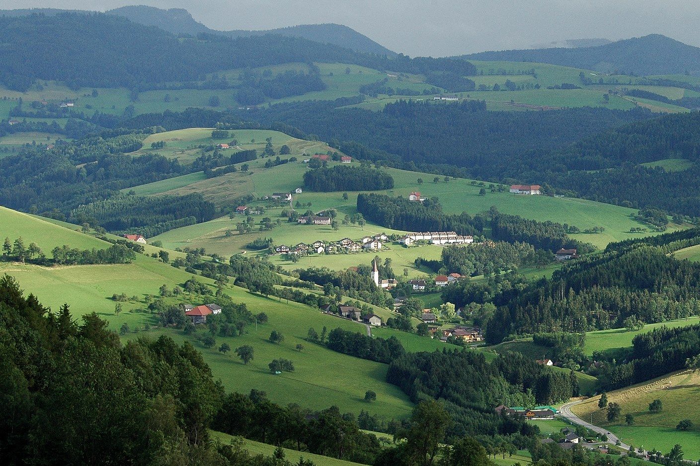 Panorámakép egy zöld, dombos tájra, elszórt házakkal és erdőkkel.