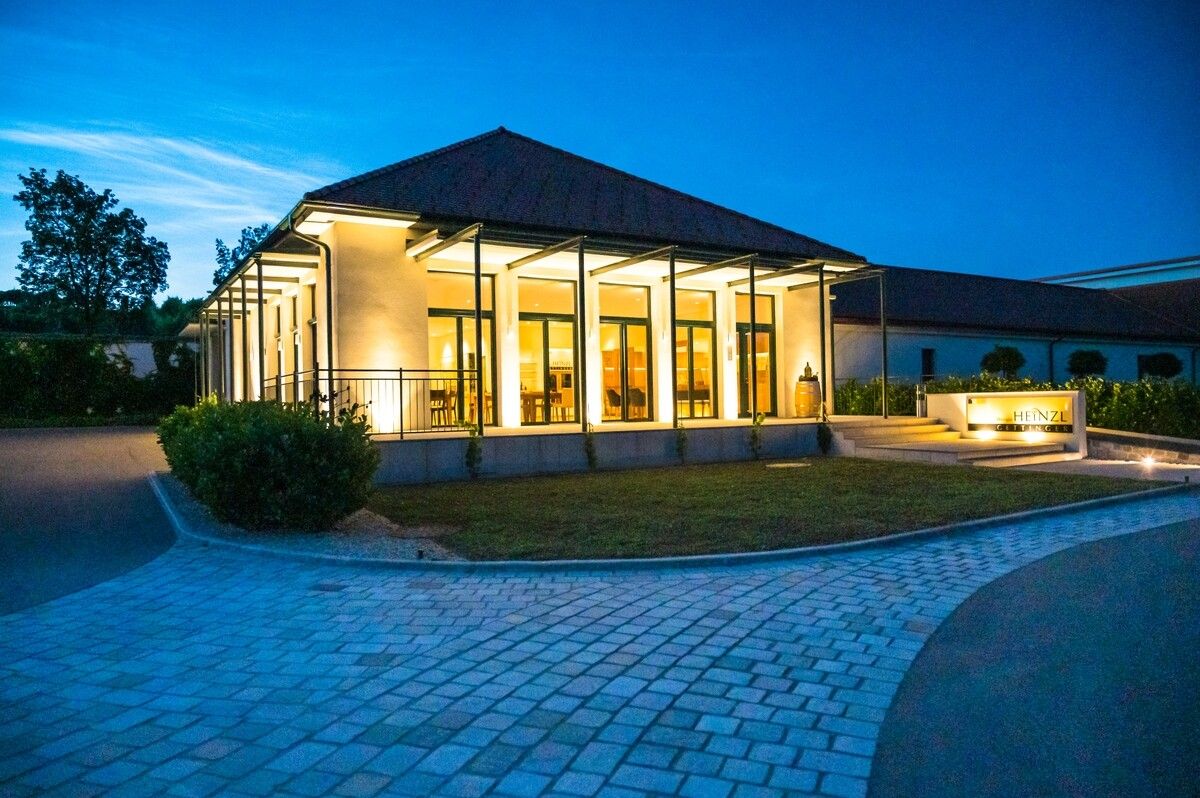 Modern épület üvegfrontokkal, szürkületben megvilágítva, burkolt út és zöldfelületek által körülvéve.