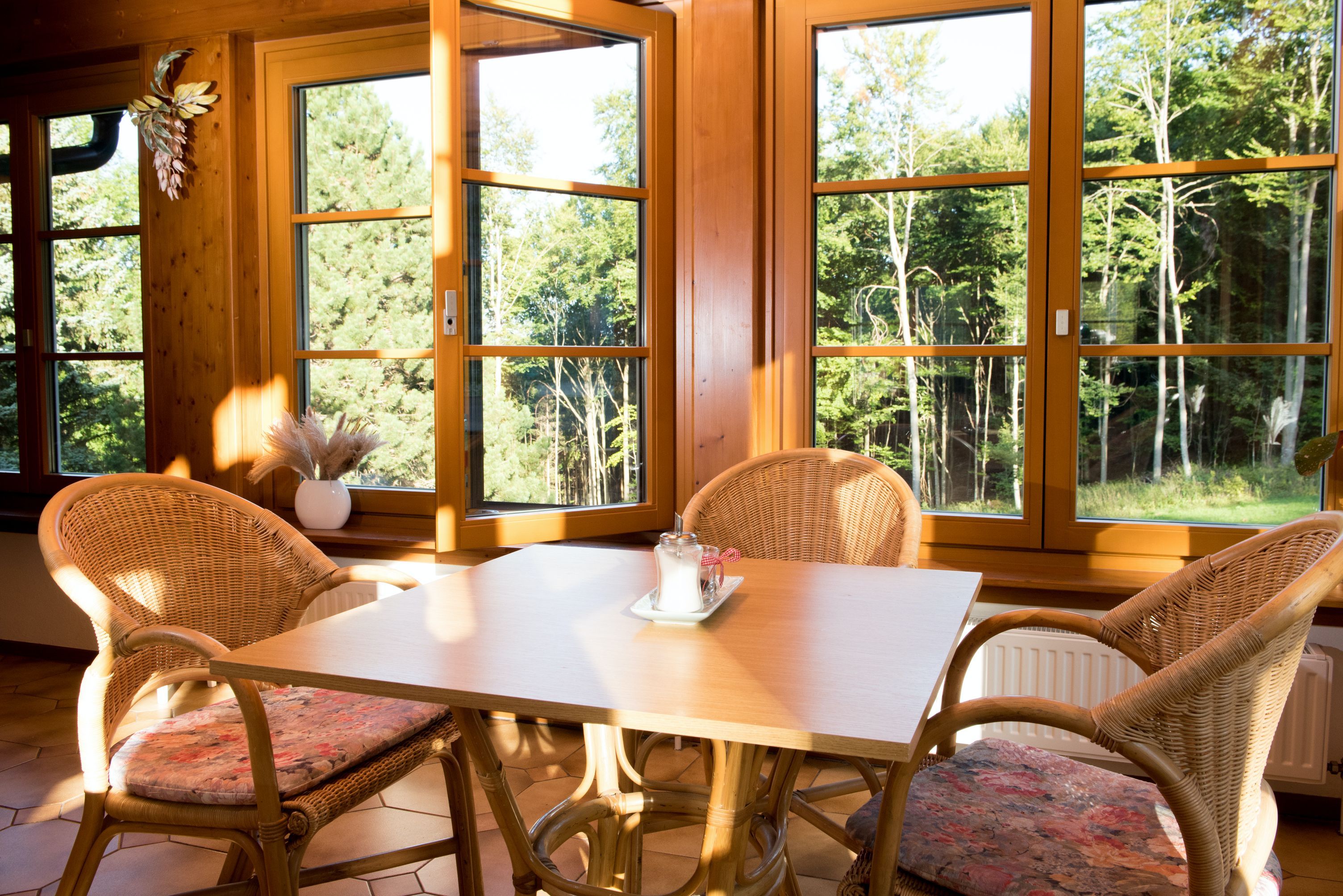 Egy asztal négy rattanszékkel egy napfényes szobában, nagy ablakokkal és kilátással az erdőre.