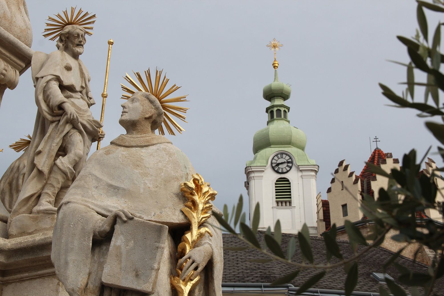 Szobor glóriával az órával és zöld tetővel ellátott templomtorony előtt.