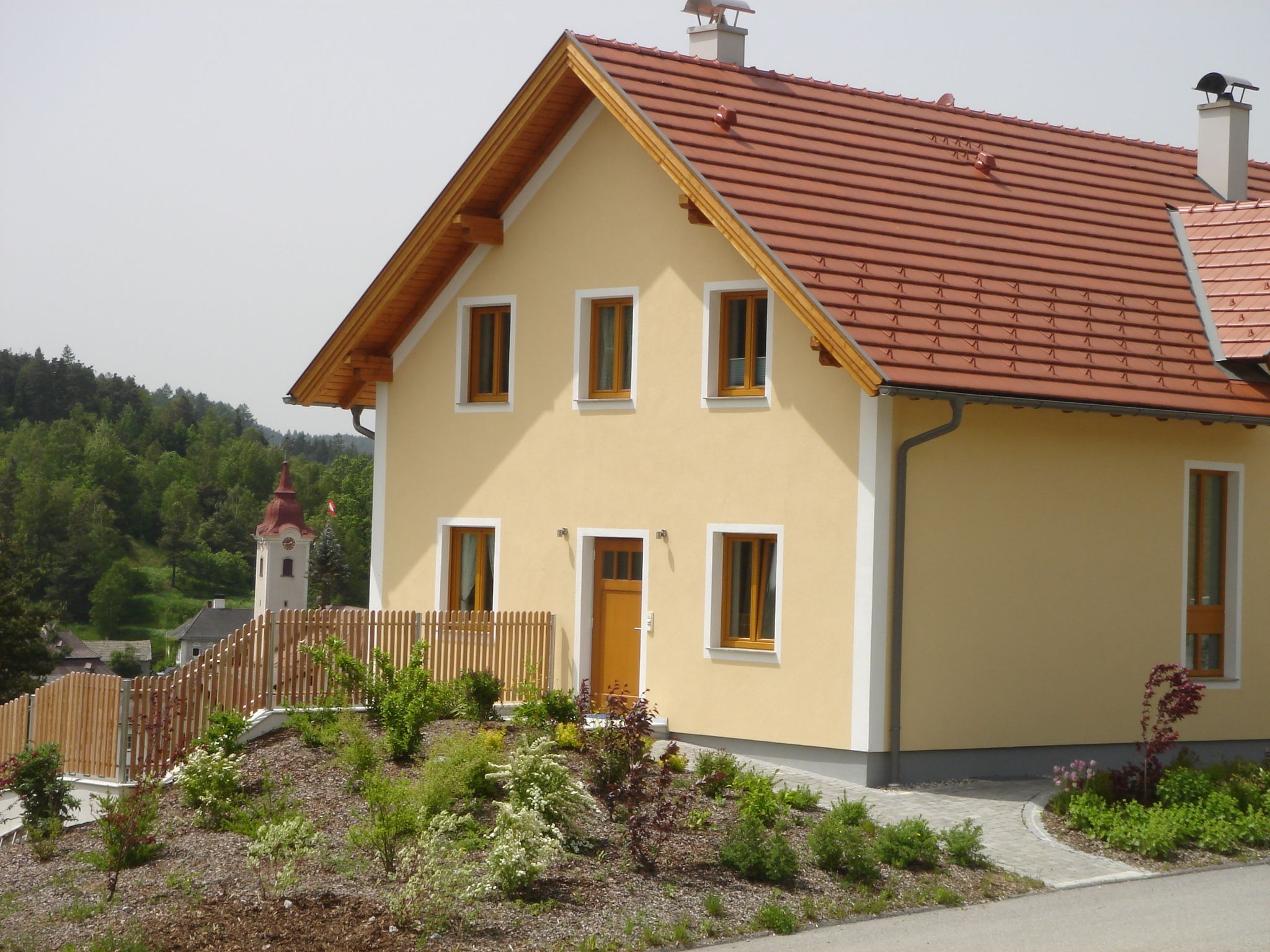 Egy modern, sárga ház piros tetővel és kerttel, a háttérben egy templommal és zöld tájjal.