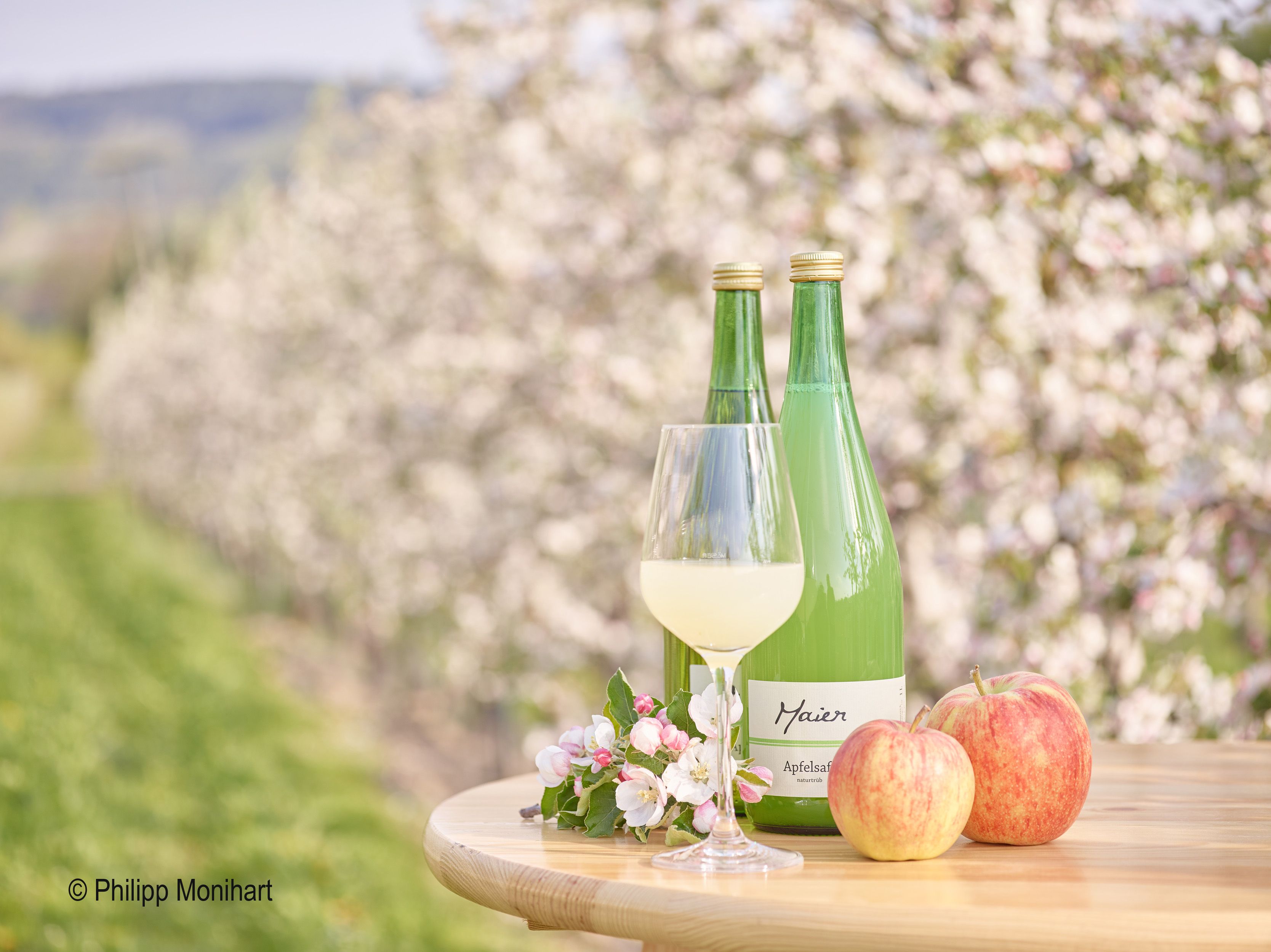 Két üveg almalé, egy pohár, két alma és almavirágok egy asztalon, virágzó almafák előtt.