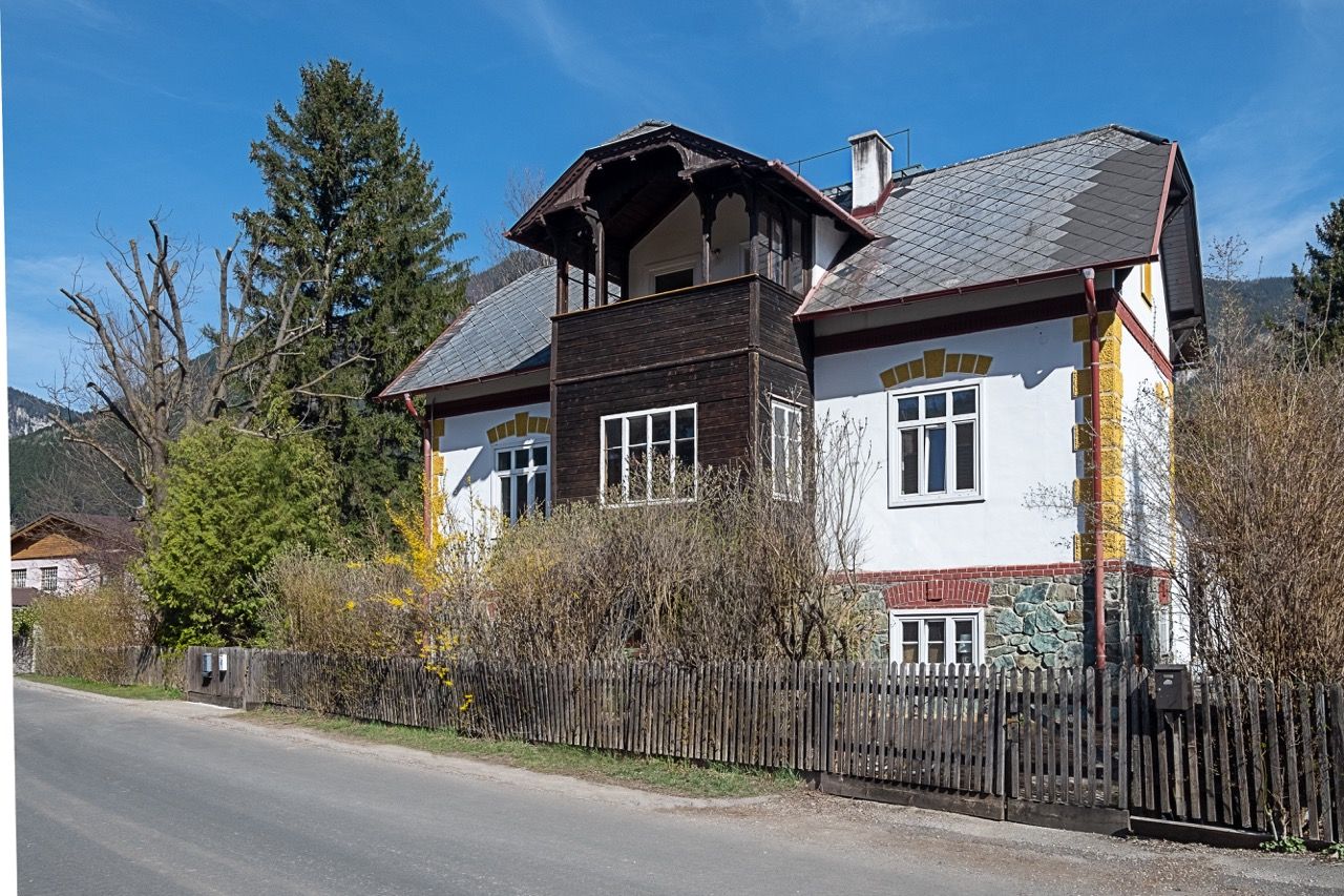 Hagyományos vidéki ház, fa erkéllyel és fehér homlokzattal, fákkal és bokrokkal körülvéve, tiszta kék ég alatt.