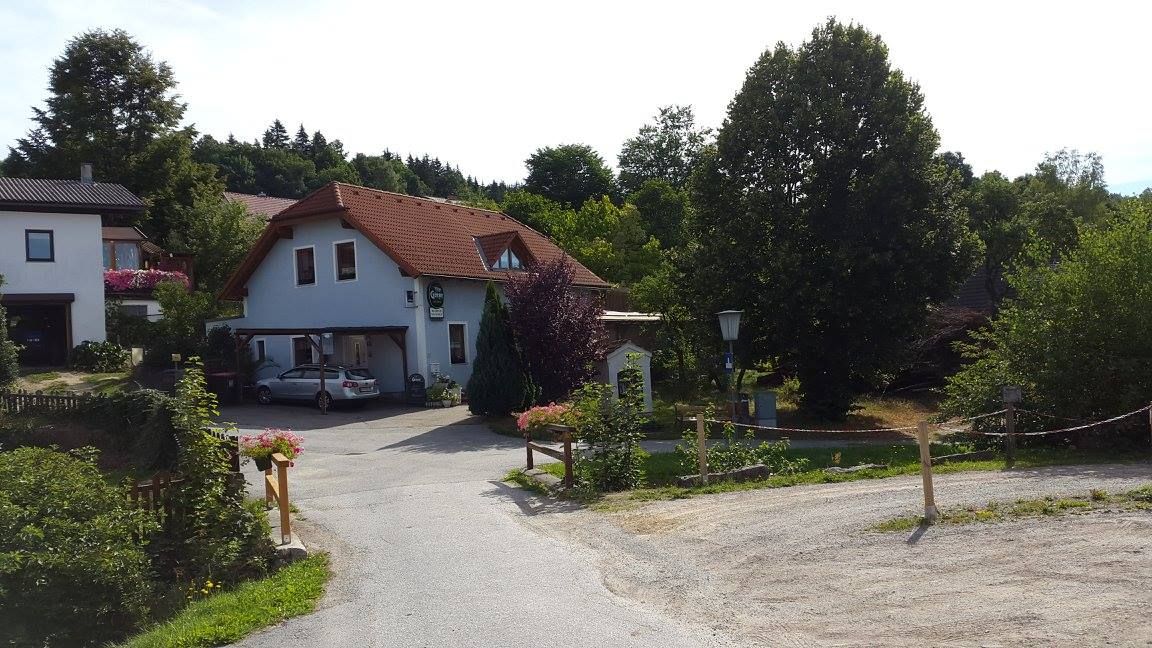 Fehér ház piros tetővel, fákkal körülvéve, az udvaron egy autó.