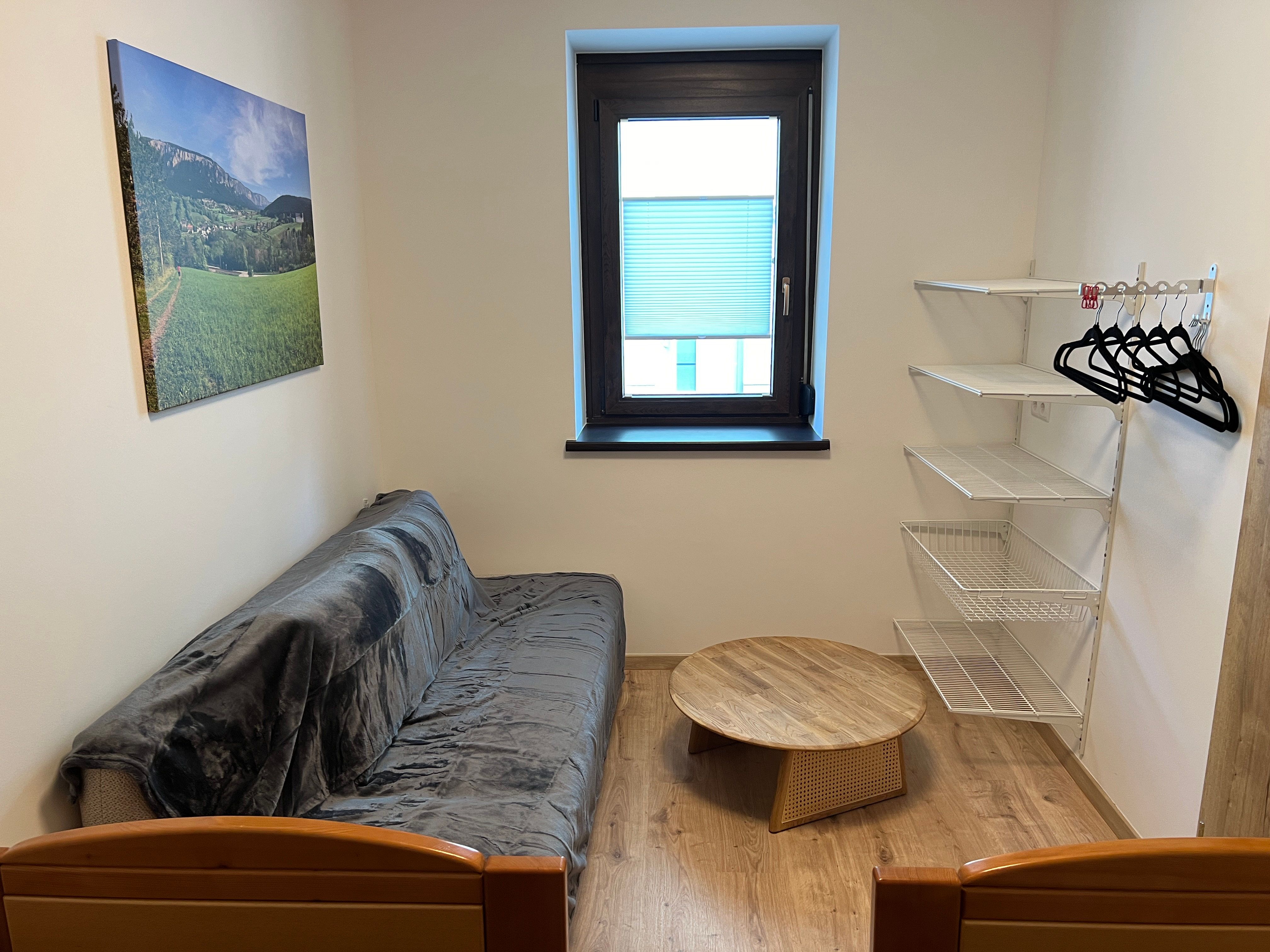 Kis szoba kanapéval, kerek faasztallal, fali polccal és ablakkal.