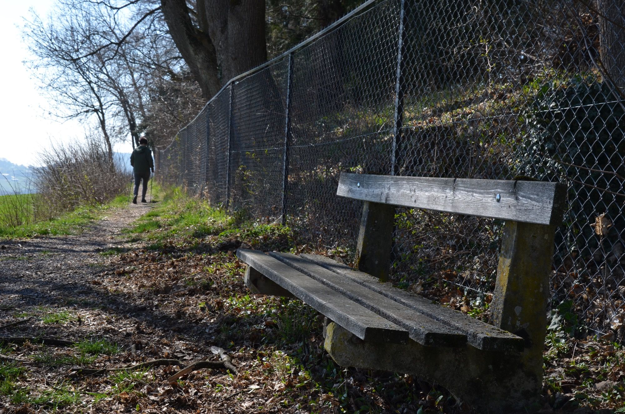 Egy gyalogösvény egy fapaddal és egy dróthálós kerítéssel. A távolban egy ember sétál.