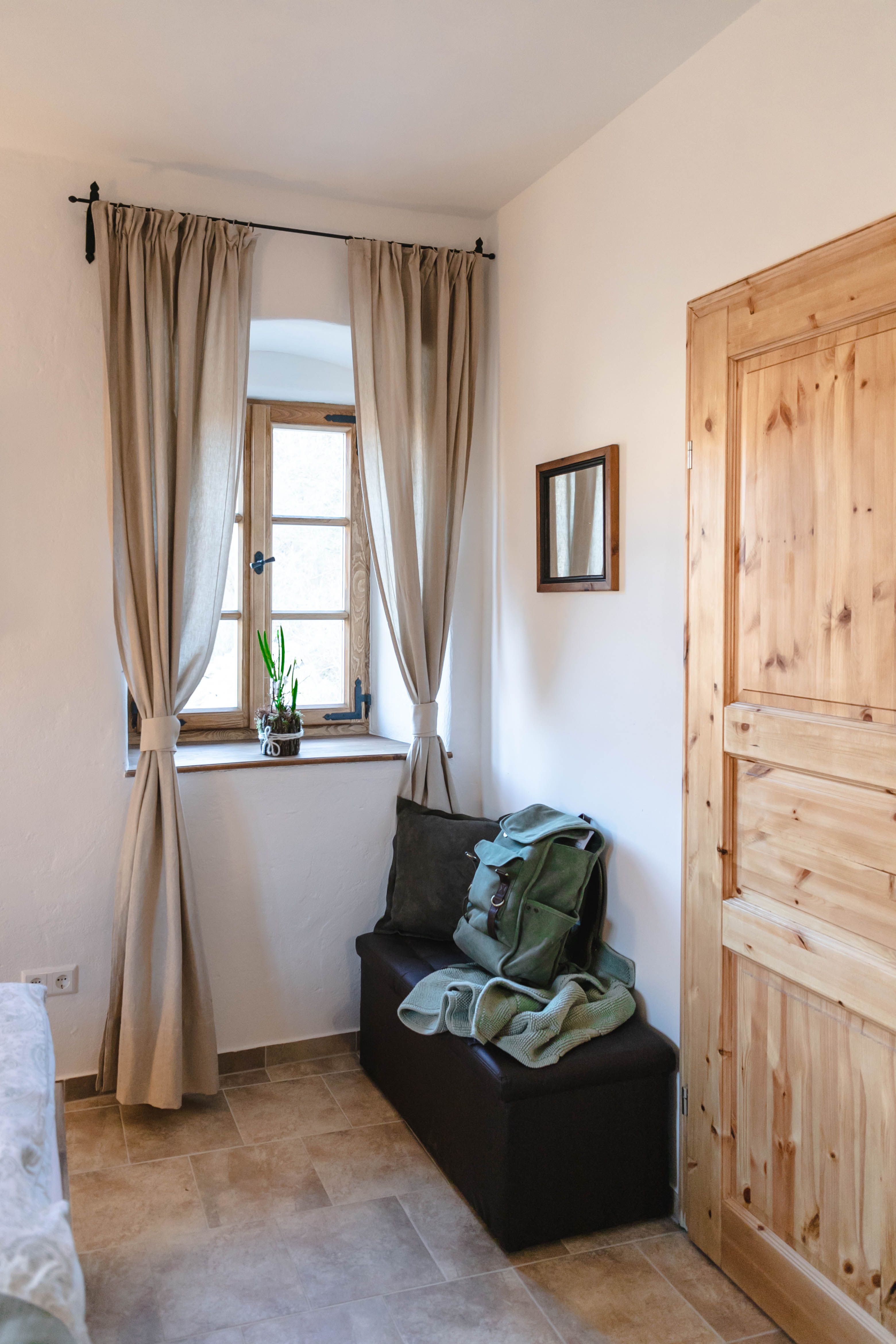 Egy kis előszoba ablakkal, függönyökkel, faajtóval és egy paddal, ahol egy hátizsák és egy takaró van.