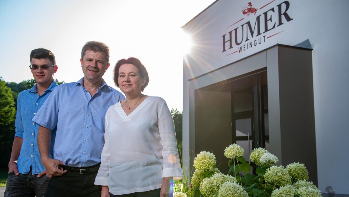 Három ember a Humer pincészet bejárata előtt, virágokkal körülvéve.