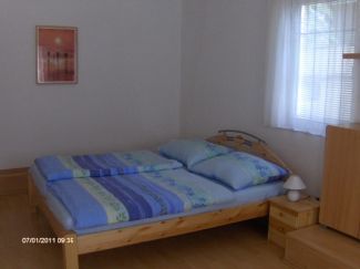 Egyszerűen berendezett hálószoba franciaággyal, éjjeliszekrénnyel és függönyös ablakkal.