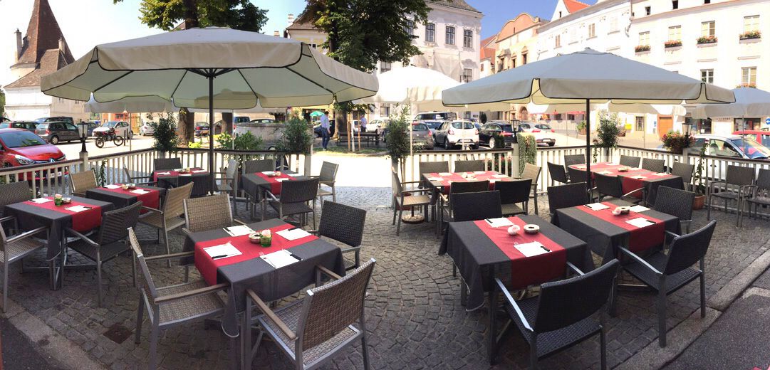 Egy kávézó szabadtéri terasza asztalokkal és napernyőkkel egy kövezett téren.