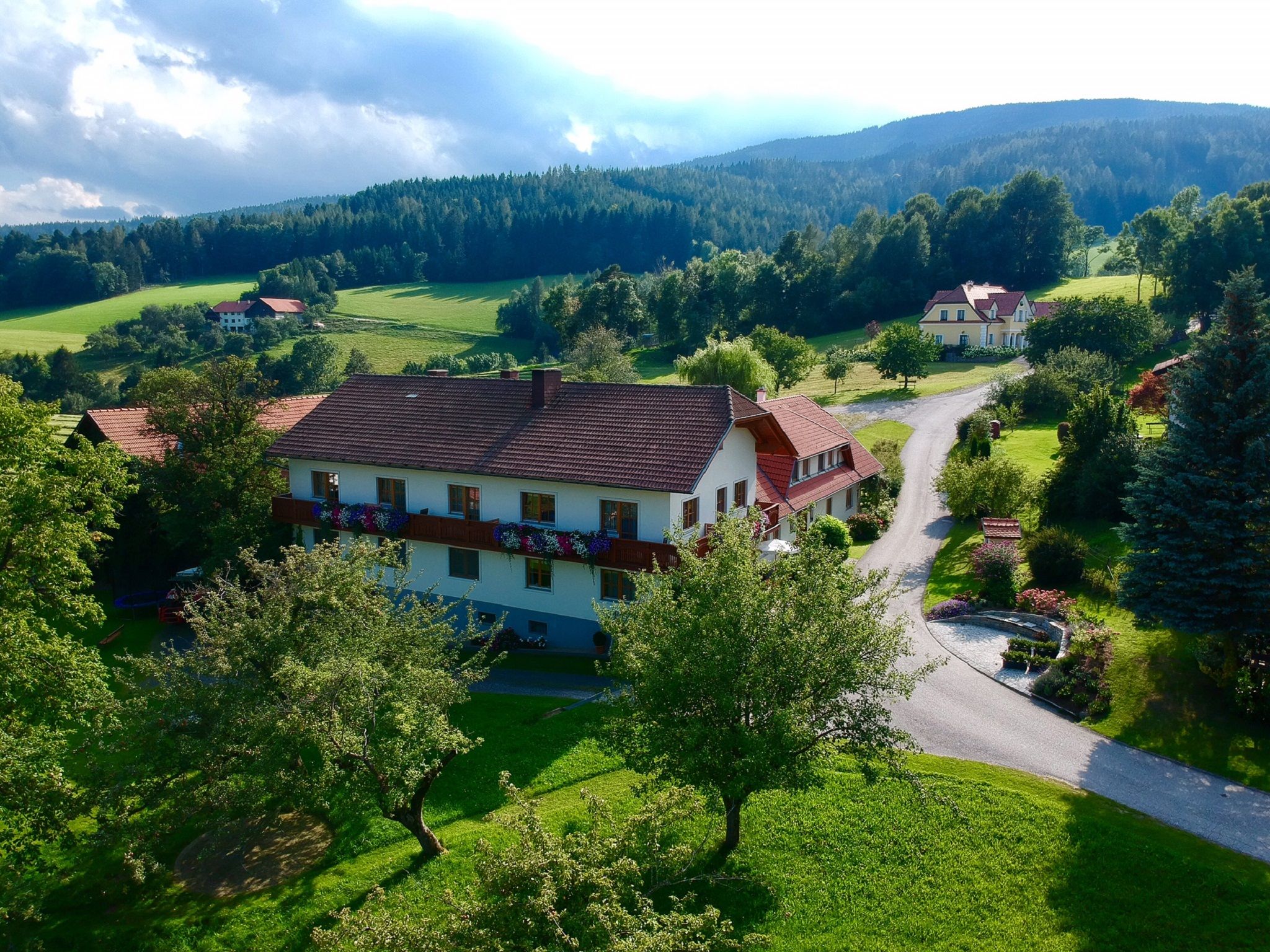 Vidéki táj egy nagy házzal, fákkal és rétekkel körülvéve, felhős ég alatt.