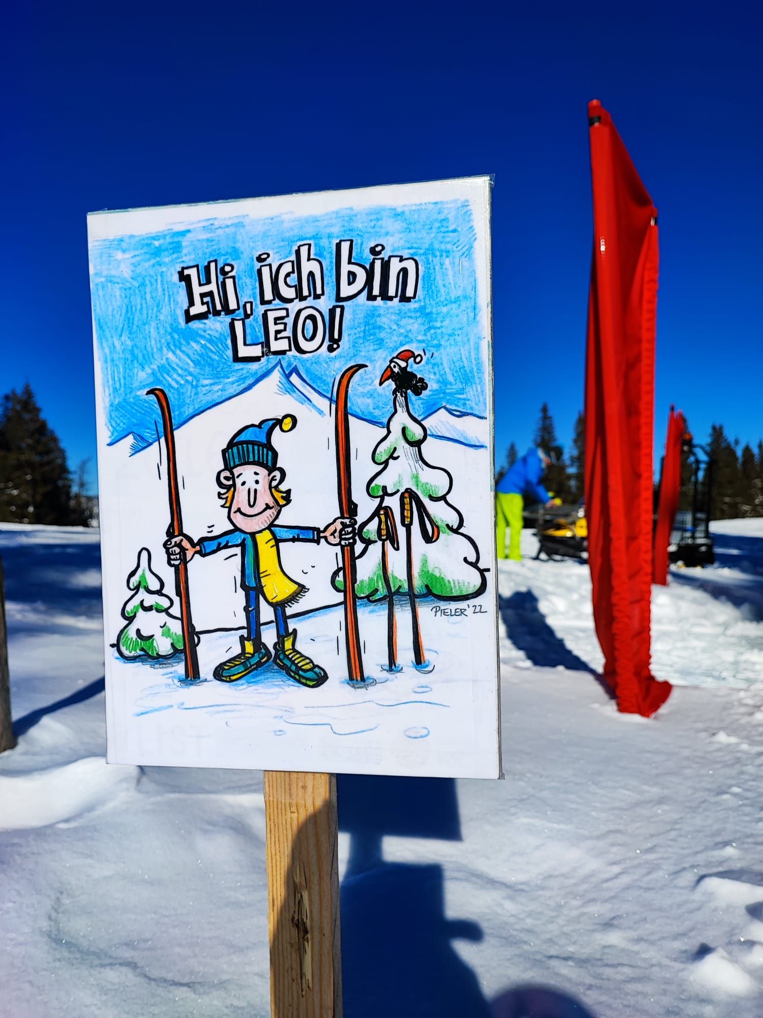 Egy tábla a hóban, rajta Leo, egy síelős rajzfilmfigura rajzával, kék égbolt előtt.