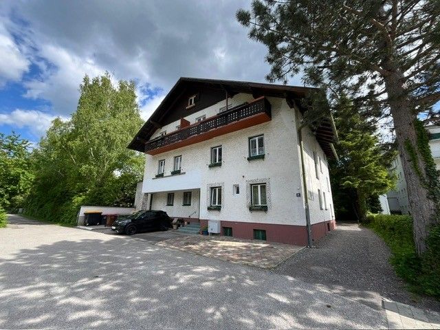 Kétemeletes ház fehér homlokzattal és sötét tetővel, fákkal és kövezett udvarral körülvéve.