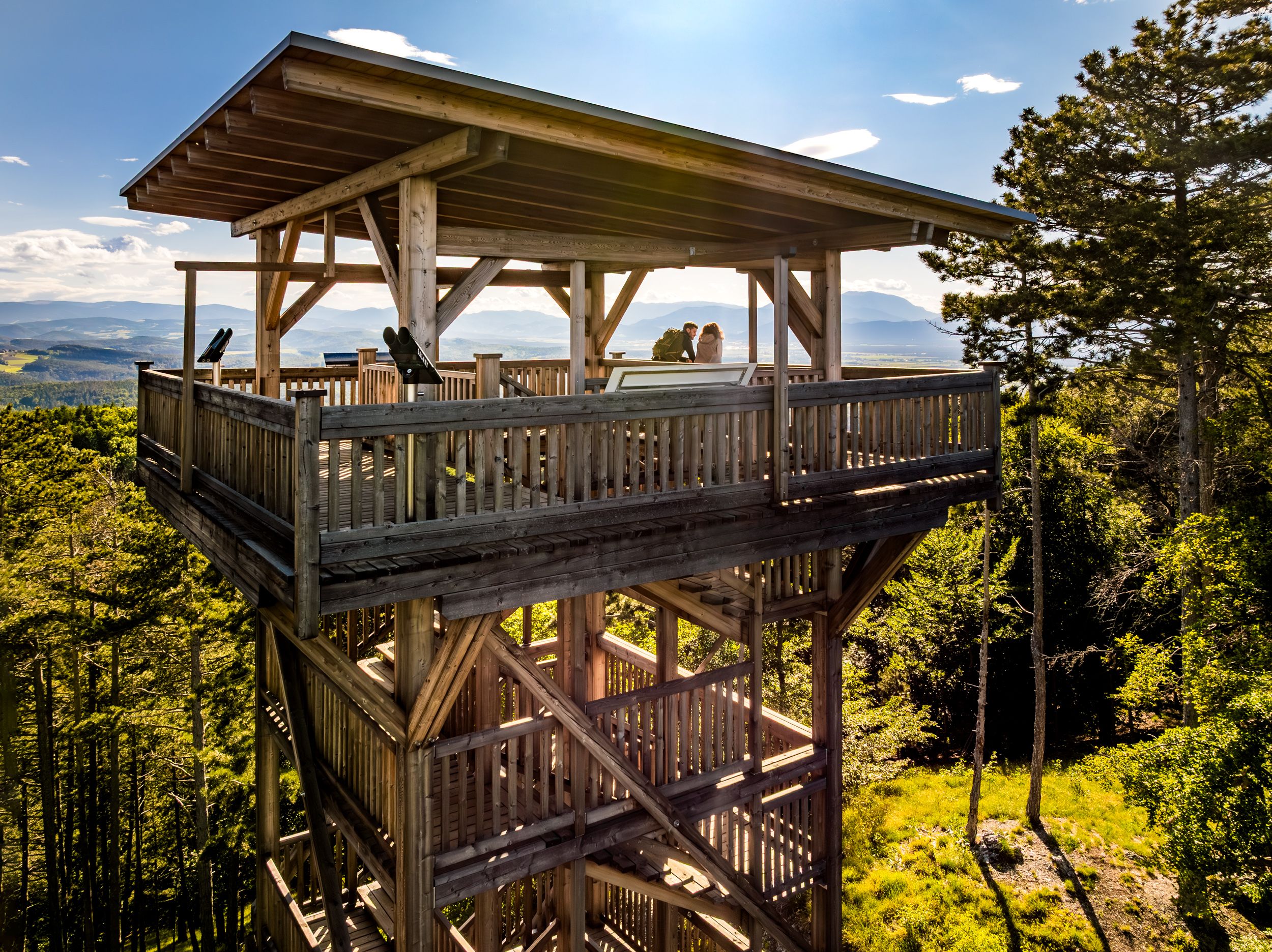 Drónfelvétel a kilátótorony oldaláról, amelyen két ember áll. A háttérben zöld erdő és hegyek