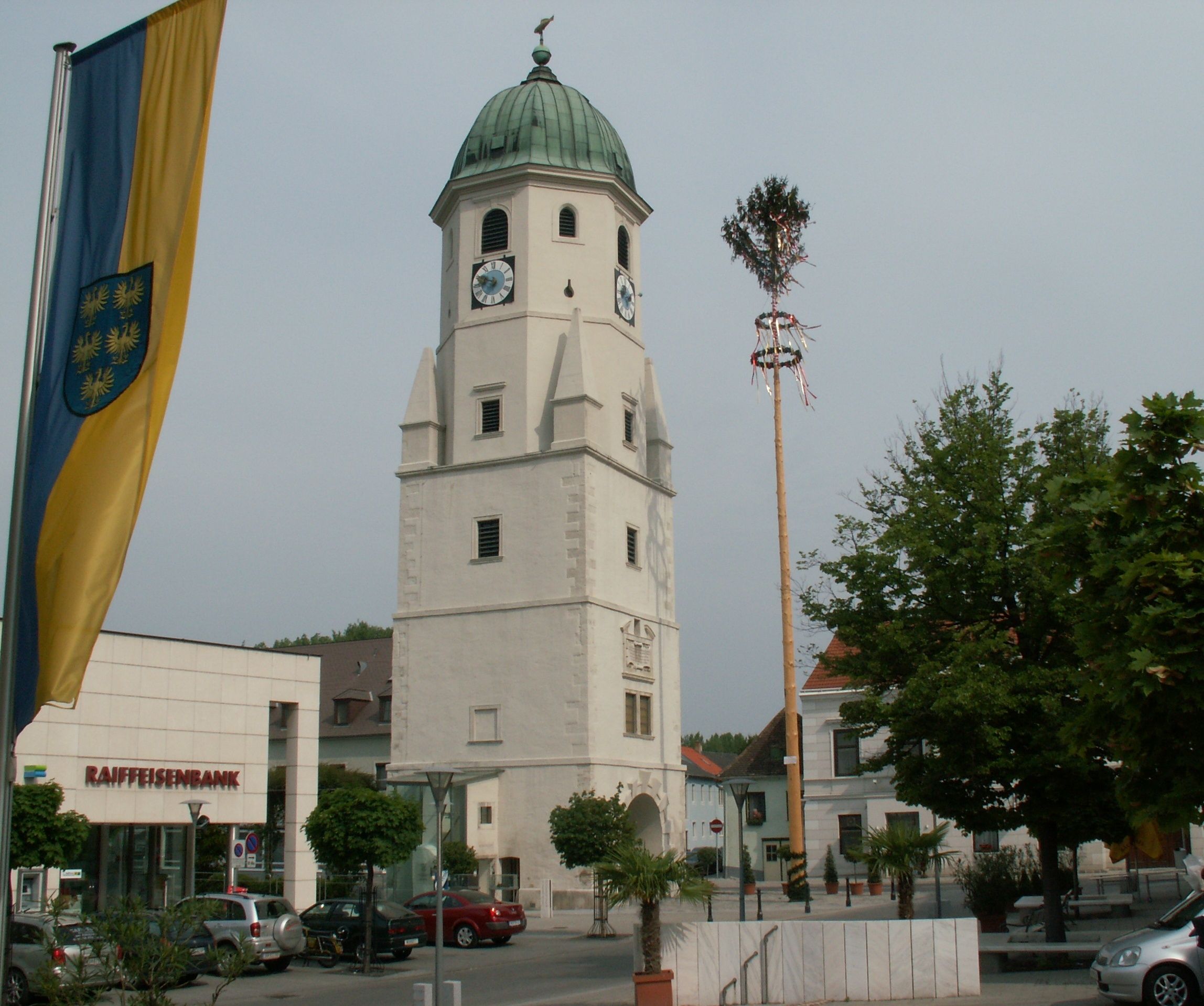 Egy templomtorony órával és zöld tetővel Fischamendben, egy májusfa és egy Raiffeisenbank fiók mellett.