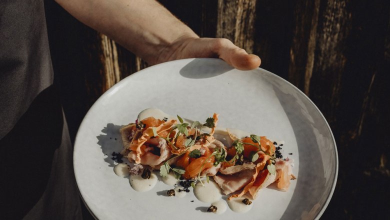 Egy tányér művészien elrendezett, fűszernövényekkel és mártással díszített vitello forello tányért nyújtanak át az egyik kézzel.